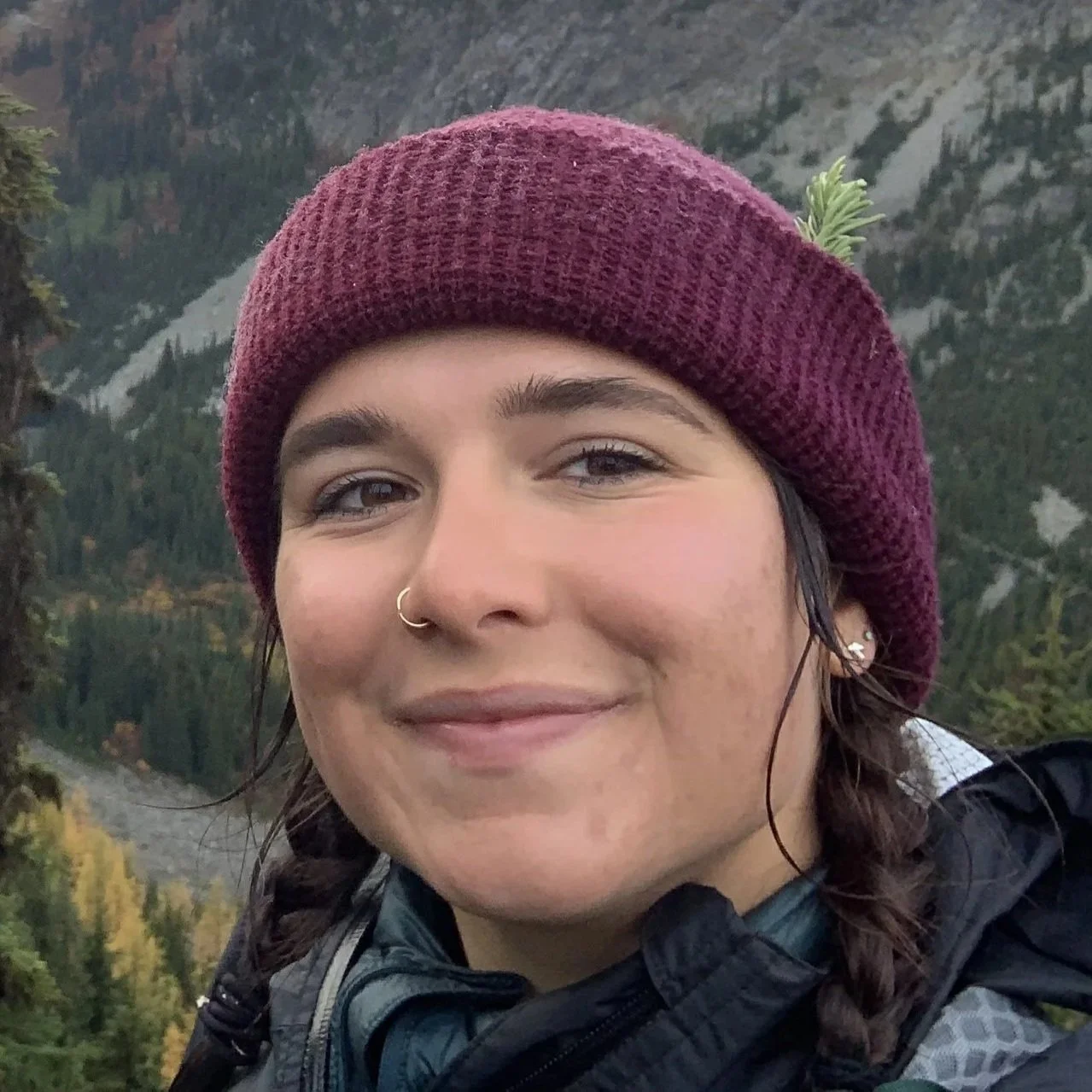 A young woman outdoors in a mountainous landscape with a lake and trees in the background, wearing a maroon knit hat and a dark jacket, smiling at the camera.
