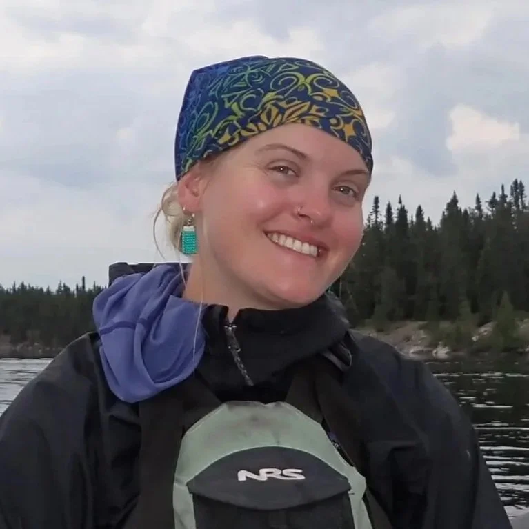 A woman wearing a colorful headscarf, black jacket, and backpack, smiling near a body of water with a forested shoreline in the background.
