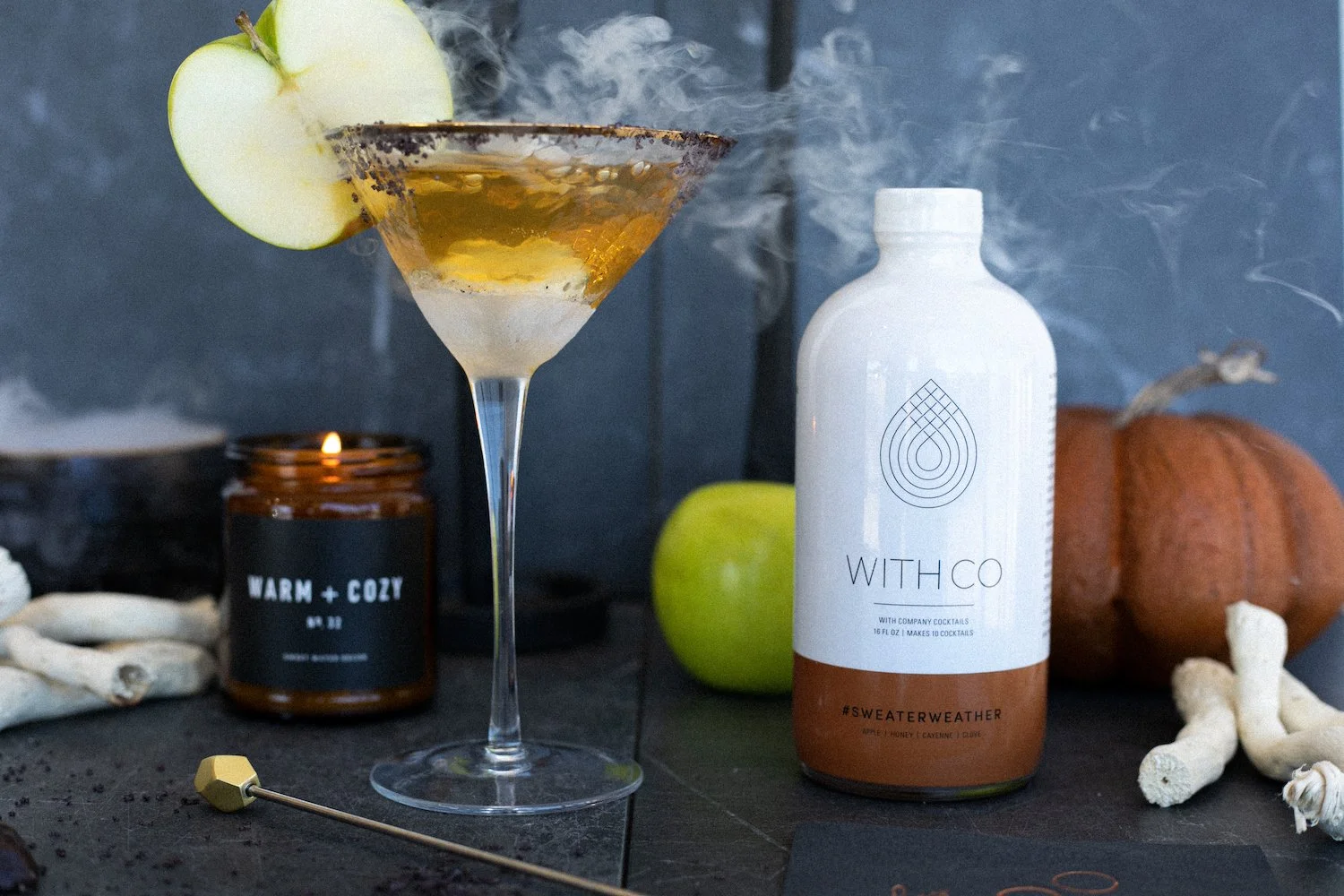 A cocktail in a martini glass garnished with an apple slice, with a bottle of WITH CO cocktail mixer, a lit candle, an apple, a pumpkin, and some decorative shells on a dark surface.