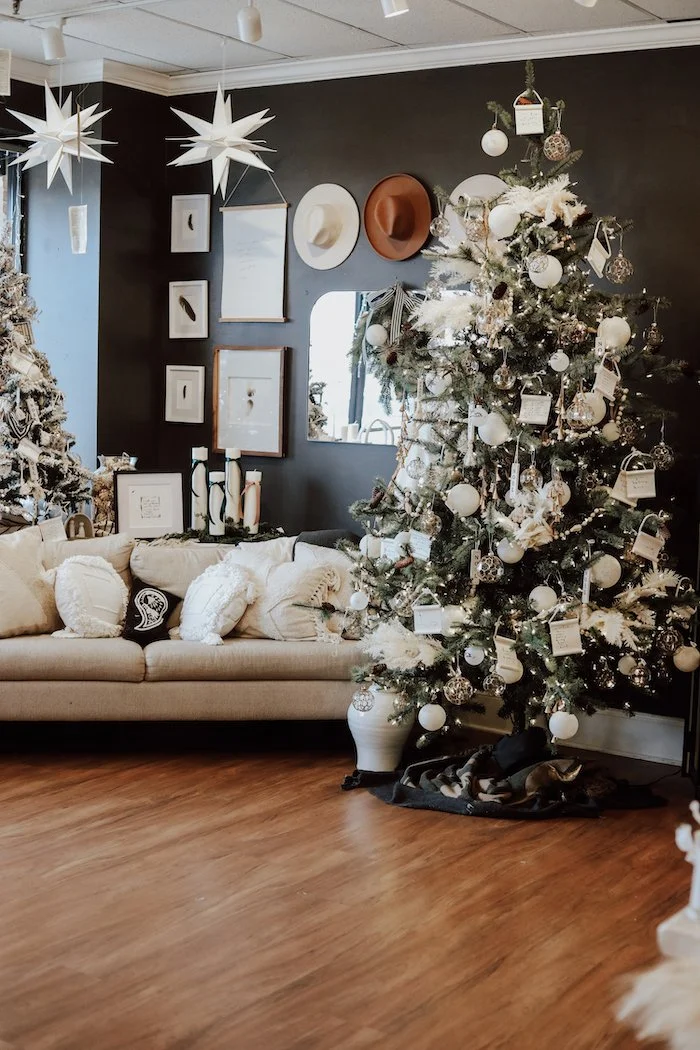 A living room decorated for Christmas features a large tree adorned with white and silver ornaments, ribbons, and tags. To the left, there is another Christmas tree with similar decorations. A sofa with beige cushions and pillows is positioned agains