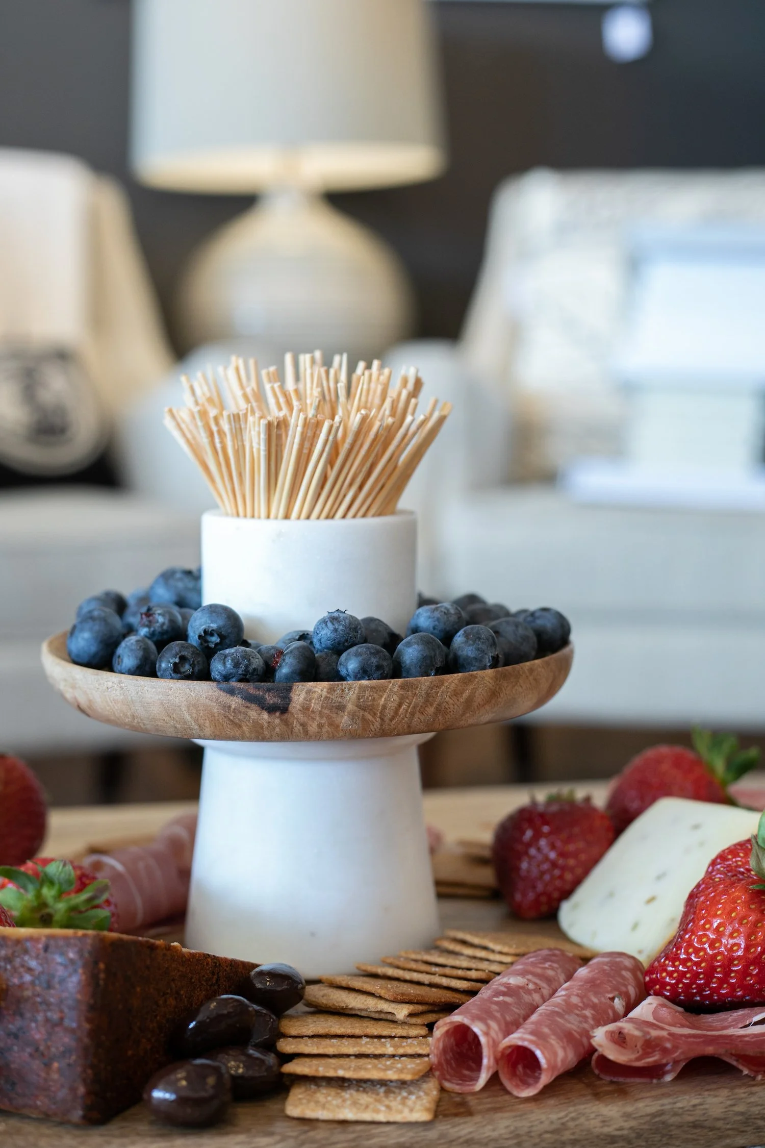 Wooden platter with blueberries, strawberries, cheese, crackers, cured meats, and black olives, with a ceramic and wooden condiment holder containing toothpicks in the center.