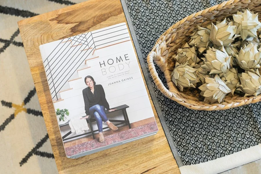 A book titled 'Homebody' by Joanna Gaines resting on a wooden table next to a woven basket filled with dried artichoke hearts.