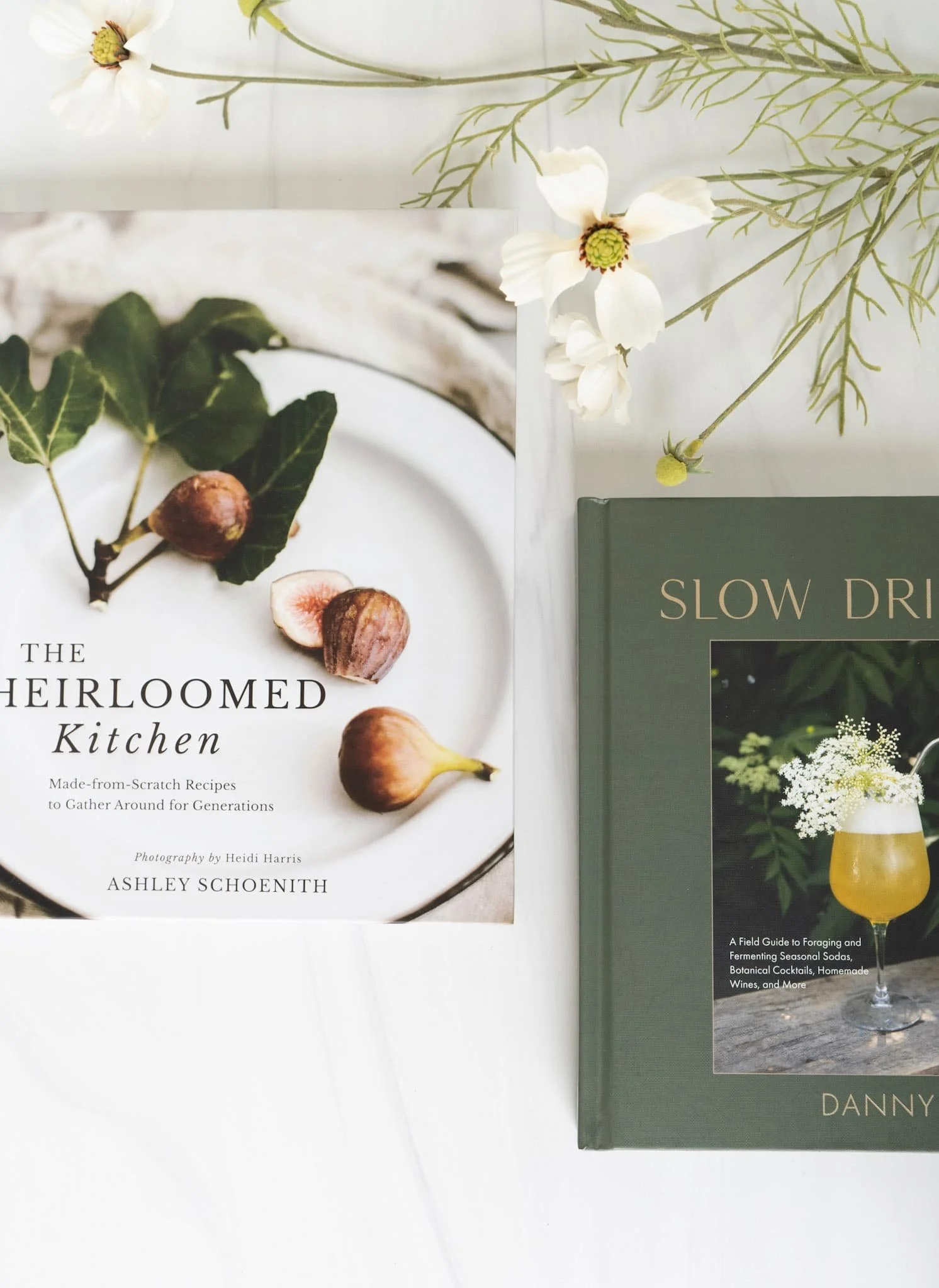 A book titled 'The Heirloom Kitchen' with figs on the cover, partly visible; next to it a green book titled 'Slow Dr' with a picture of a drink on the cover; white flowers and greenery arranged decoratively on a white surface.