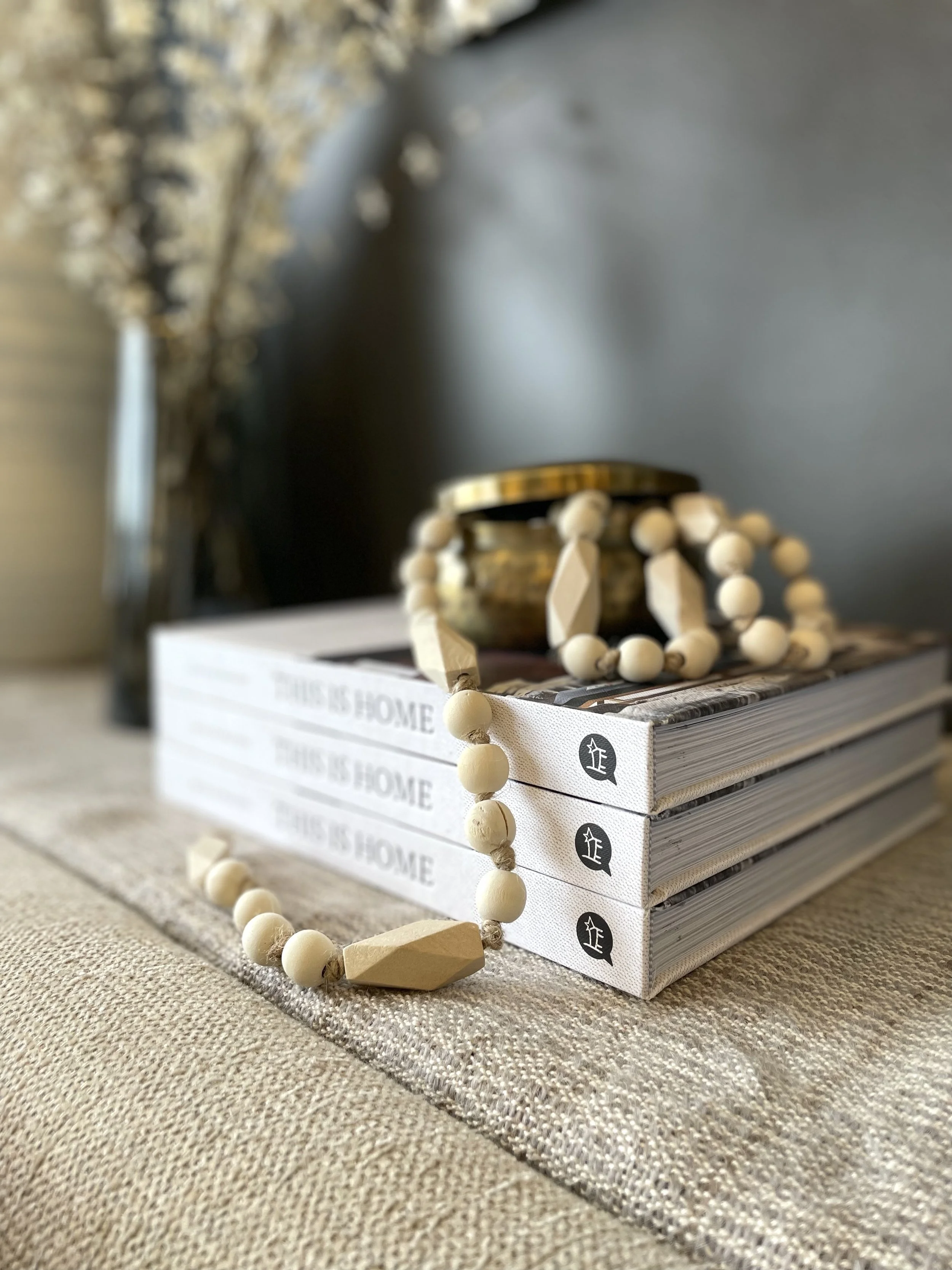 Decorative objects including a stack of three books with "THIS IS HOME" printed on the spines, a wooden bead necklace draped over the books, a small brass container, and a blurred background with a potted plant on the left.