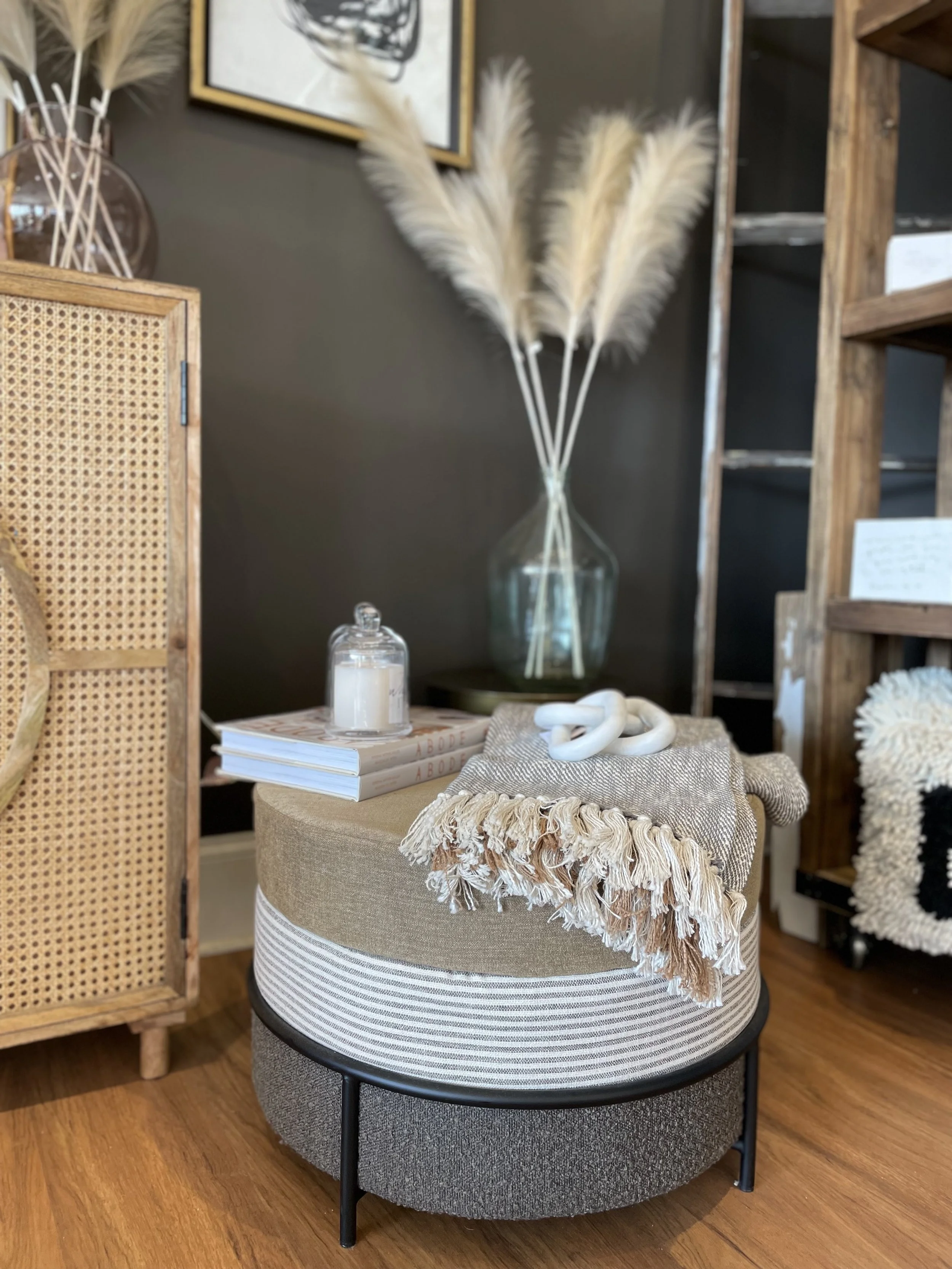 Decorative home interior with a glass vase of pampas grass, a candle on top of books, and a beige fringed throw blanket on a round ottoman.