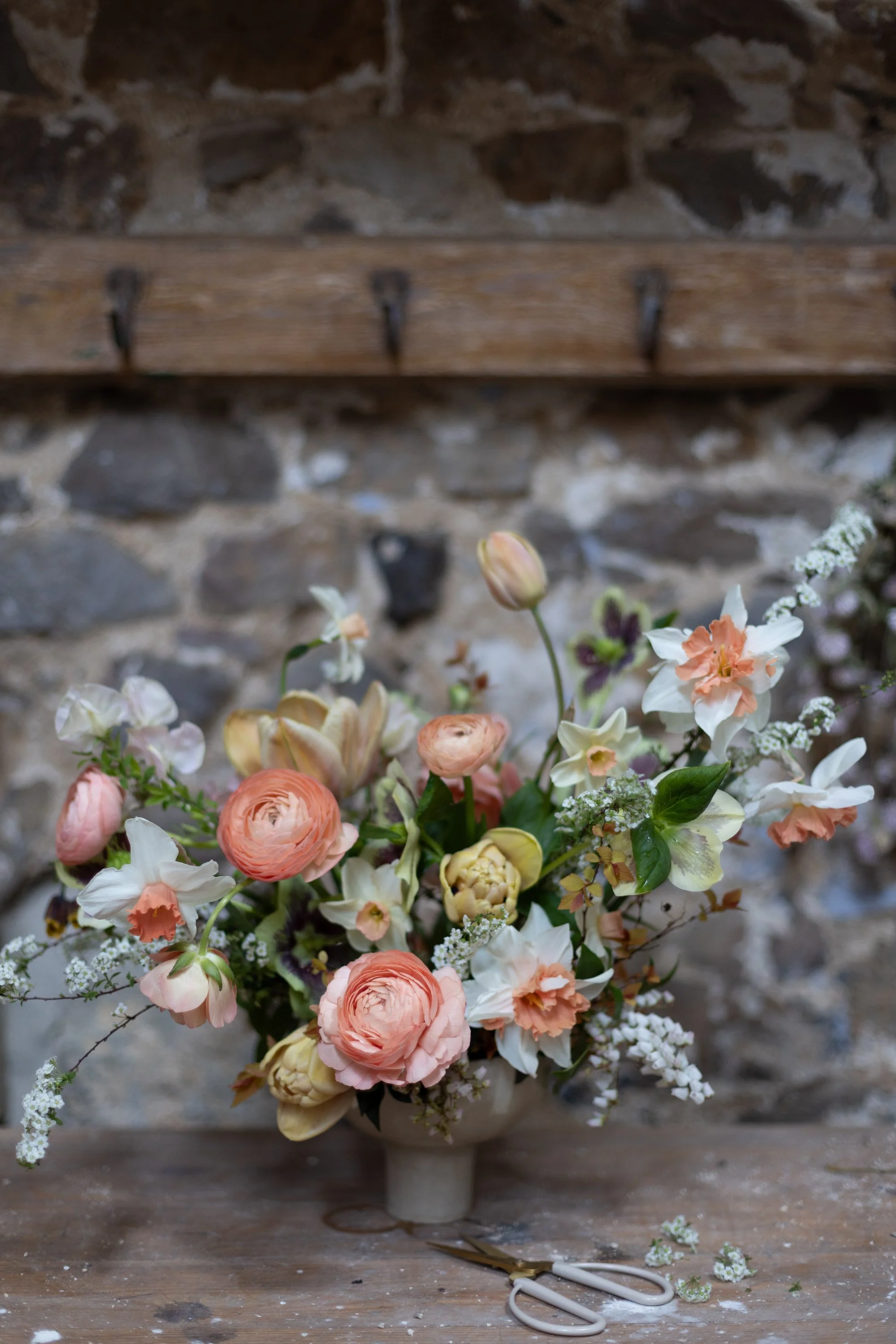 A bouquet of pink, white, and peach flowers arranged in a vase on a rustic wooden table, with garden scissors and scattered small white flowers nearby.