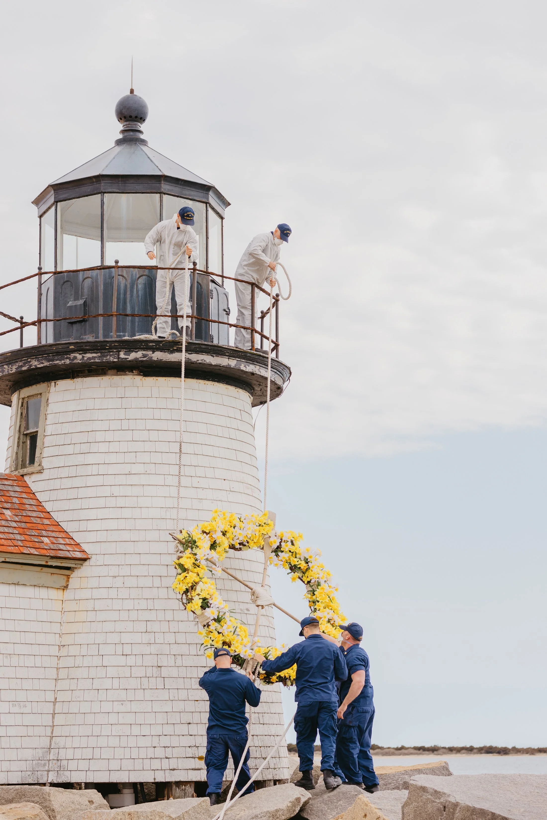 Six men in uniform are hanging a wreath decorated with yellow and white flowers on a lighthouse. Two of the men on the lighthouse are working with ropes, one is holding the wreath, and the others are assisting at ground level.