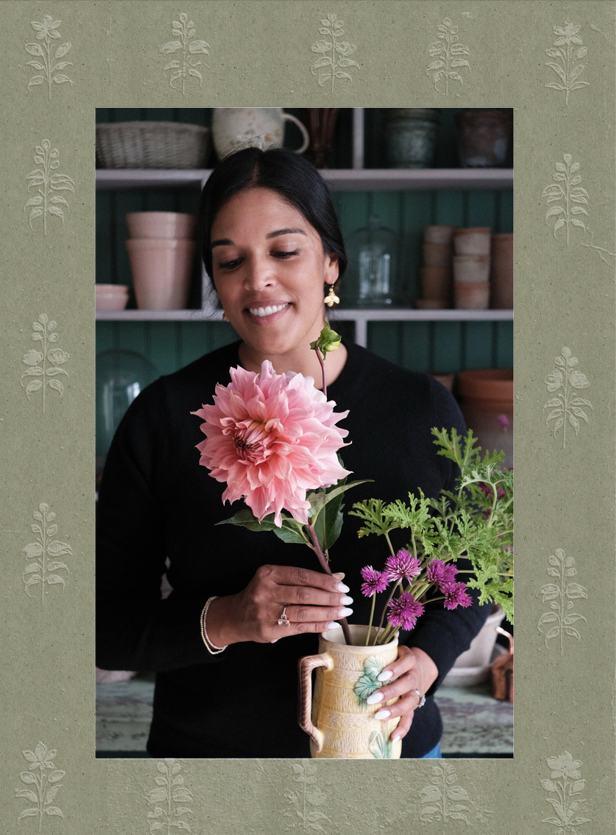 A woman holding a vase filled with pink, purple, and green flowers, standing indoors with shelves of pottery in the background.