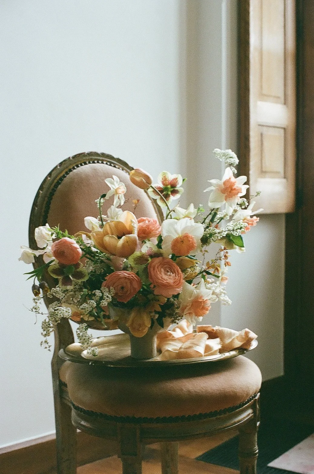 A bouquet of pink, white, and cream flowers in a vase sitting on a vintage chair with a beige cushion, in a softly lit room with a window and wooden shutters in the background.