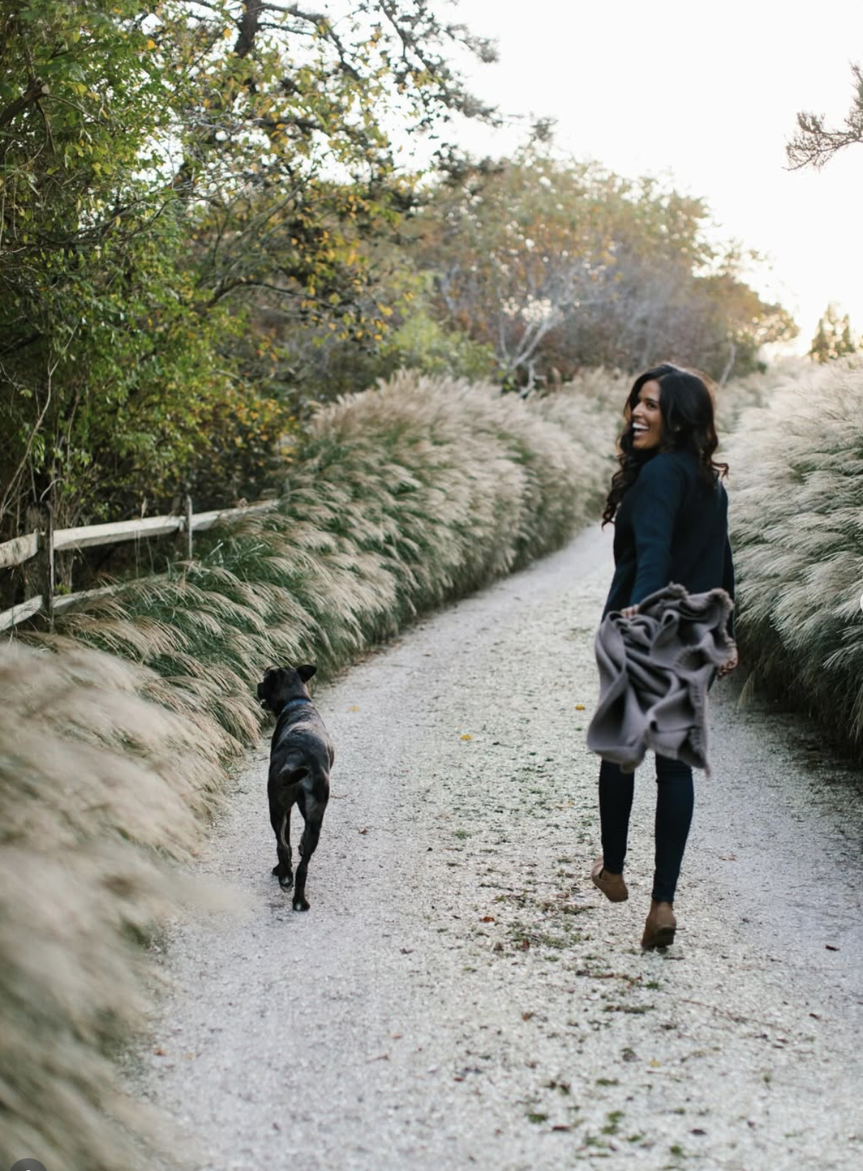 A woman walking on a dirt path with her dog during daytime. She is smiling and holding a coat, with trees and tall grasses on both sides of the path.