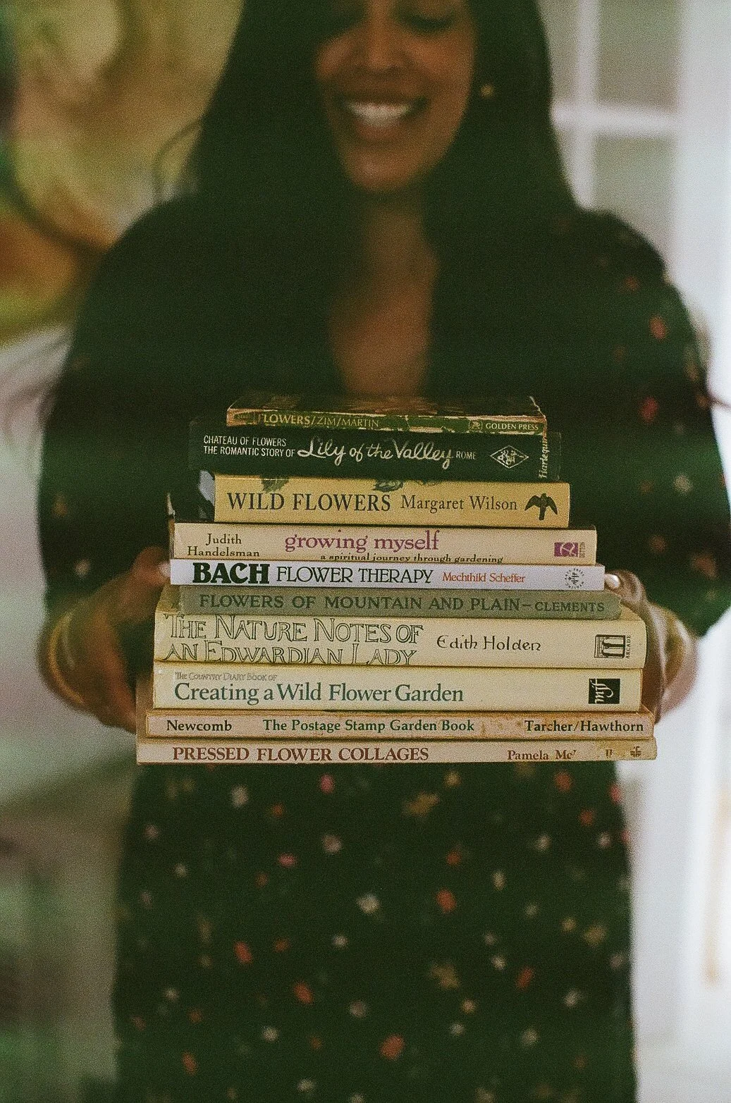 A woman with dark hair and a floral dress is holding a stack of books about flowers and gardening. She is smiling and standing indoors.