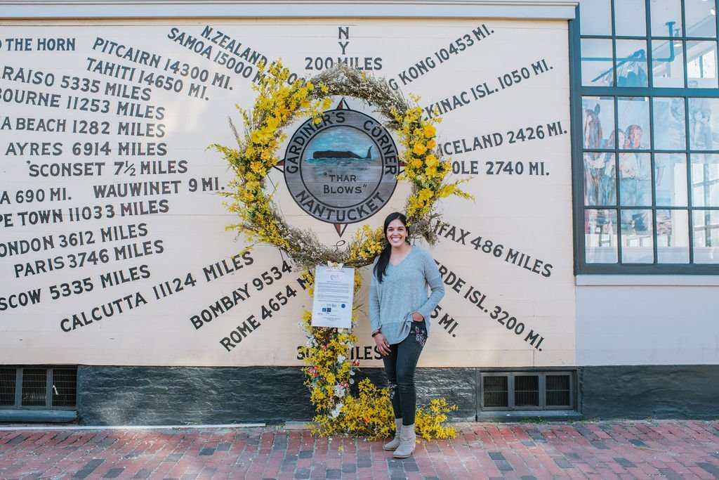 Woman standing next to a decorative wreath with a sign at Gardner's Corner, Nantucket, featuring a wooden sign and a wall with various world distances.