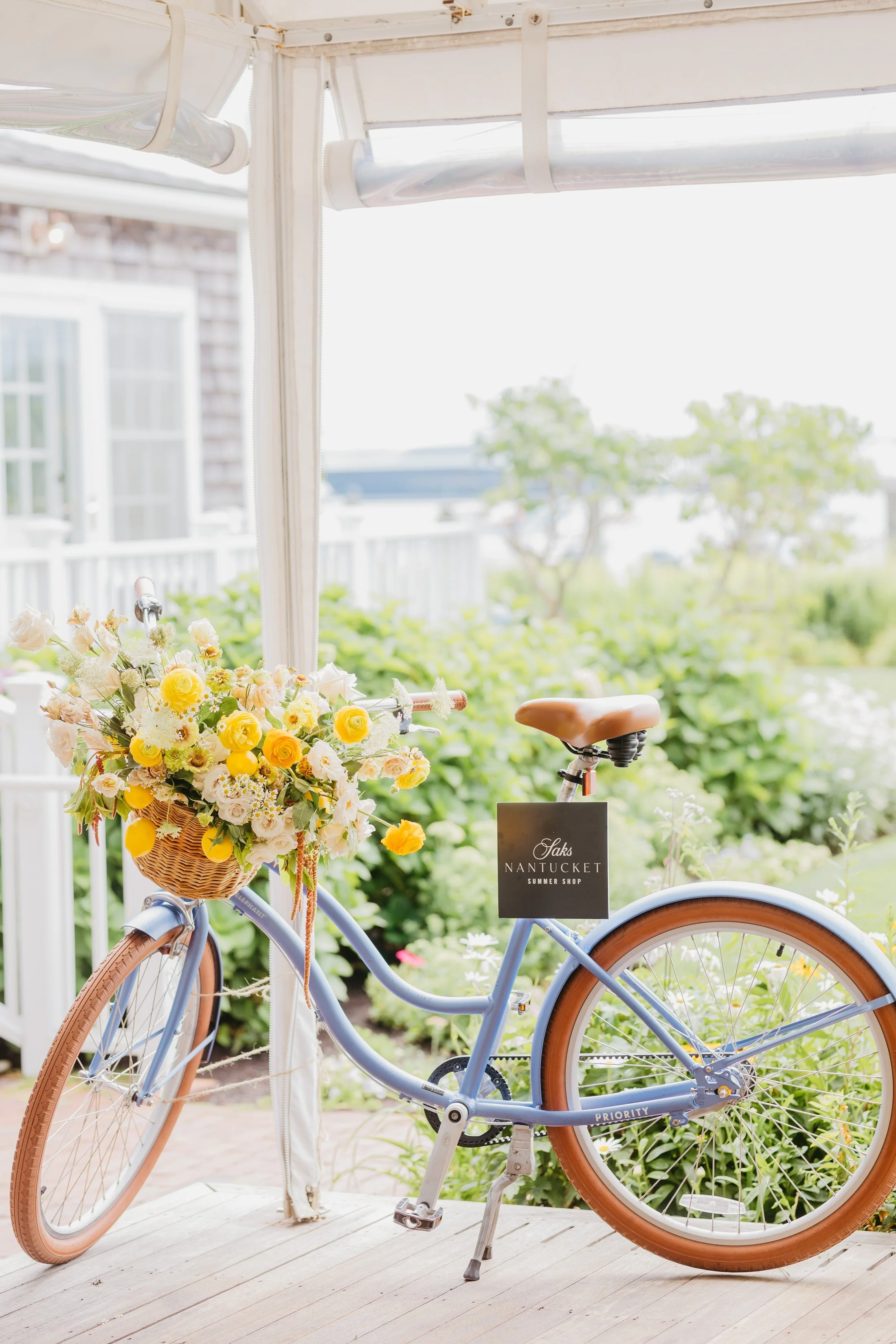 A light blue bicycle with brown tires and a basket filled with yellow and white flowers, parked on a porch with greenery in the background, and a black sign that reads 'Oaks Nantucket Summer Shop' attached to the bike.