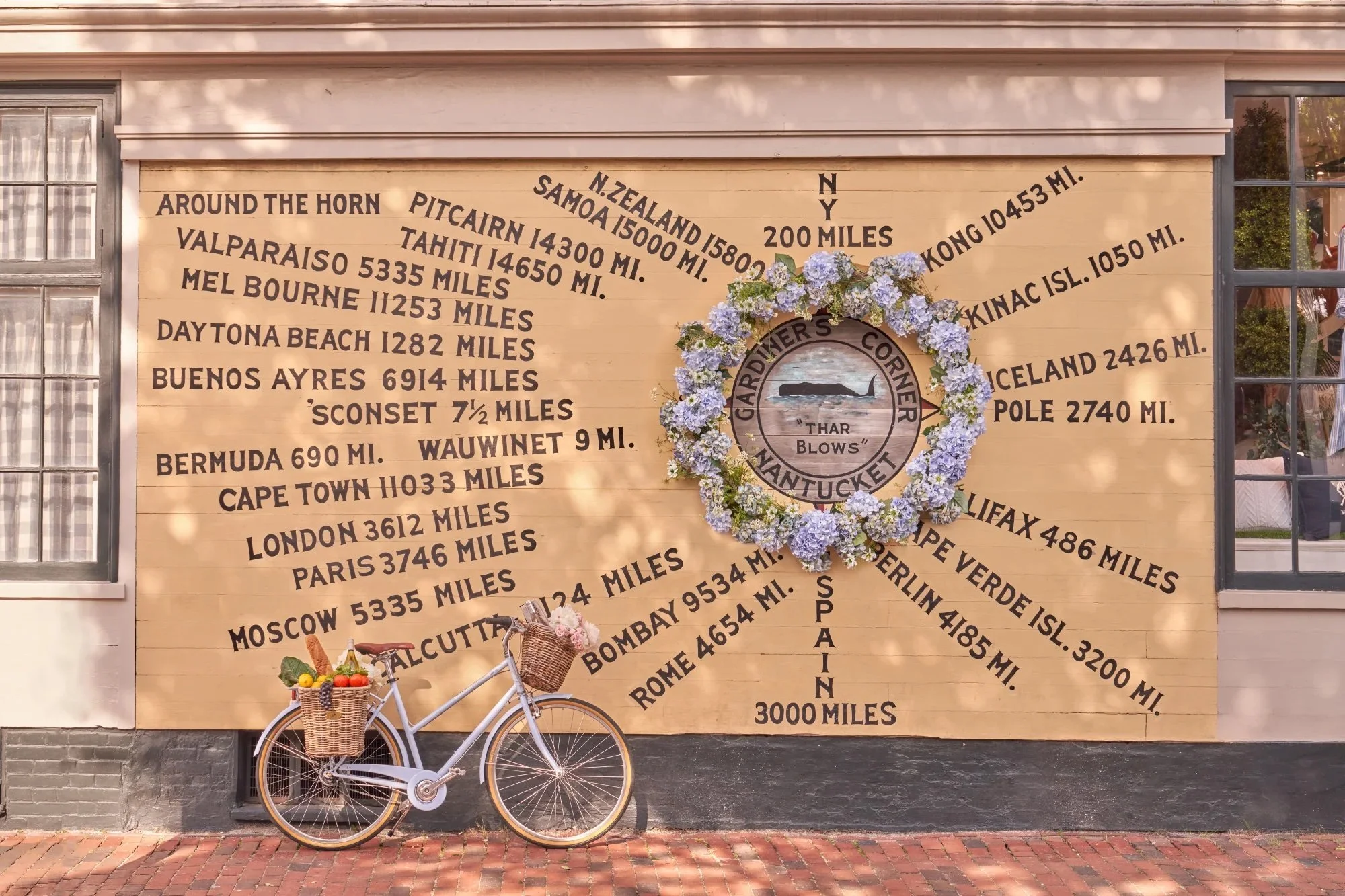 A wall with a list of long-distance travel distances in miles, arranged in a semi-circular pattern, surrounded by a flower wreath, with a bicycle decorated with a basket of fruits and flowers in front of it.