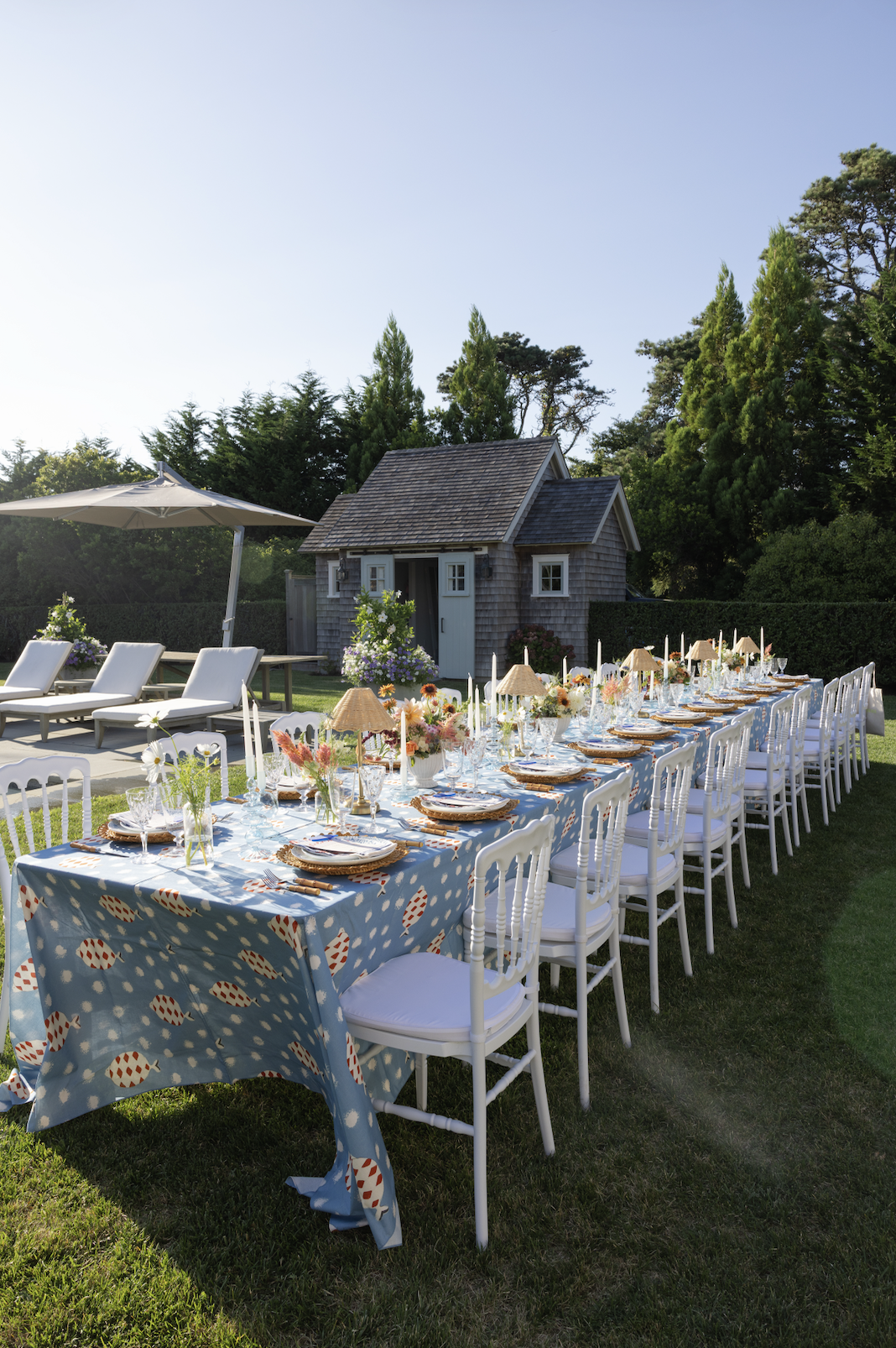 Long outdoor dining table set with white chairs, decorated with colorful flowers, candles, and tableware, in a backyard with a small wooden house, lounge chairs, umbrella, and lush green trees in the background.