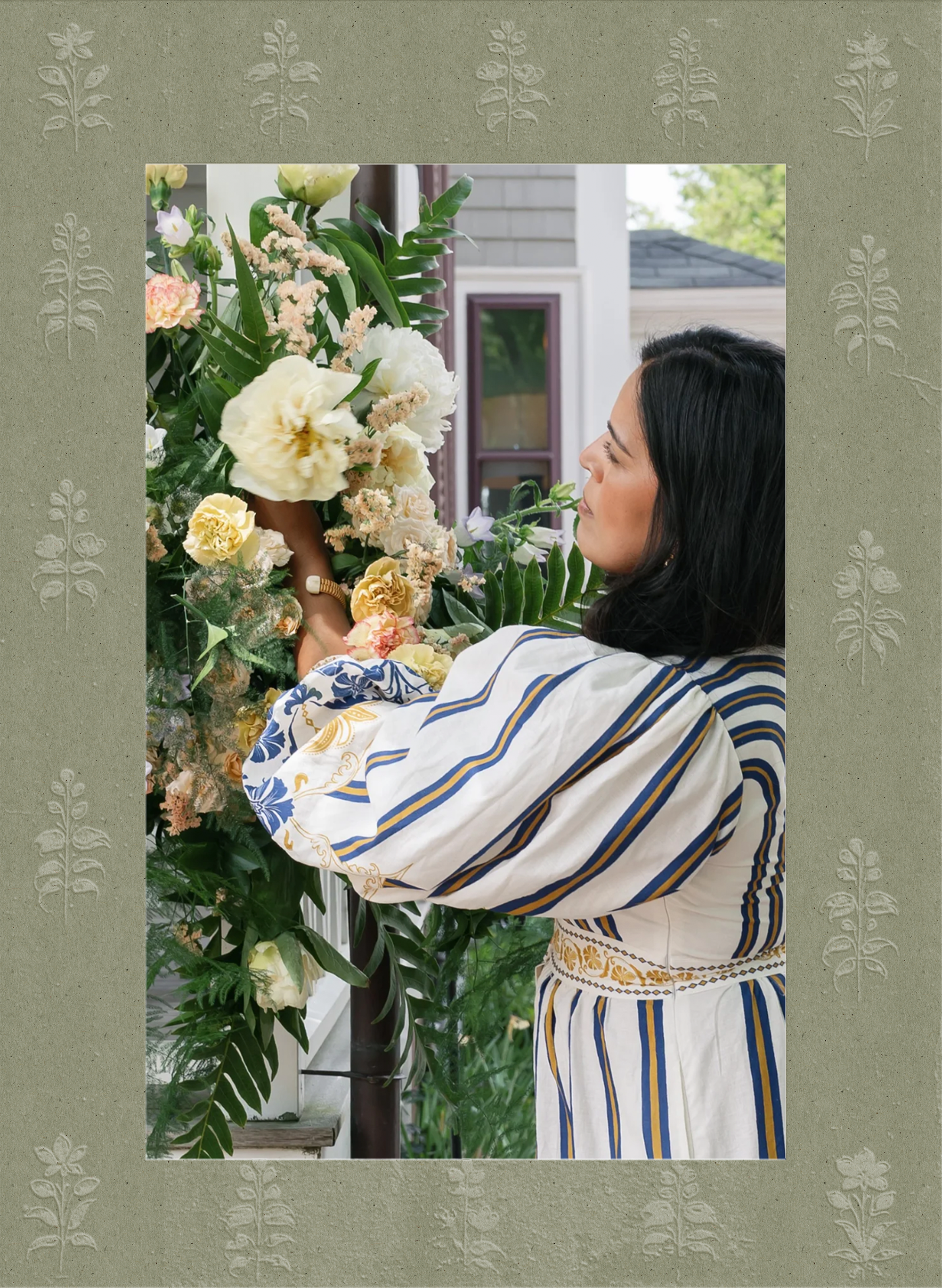 Woman arranging a large bouquet of flowers on a porch or patio, with a house and greenery in the background.