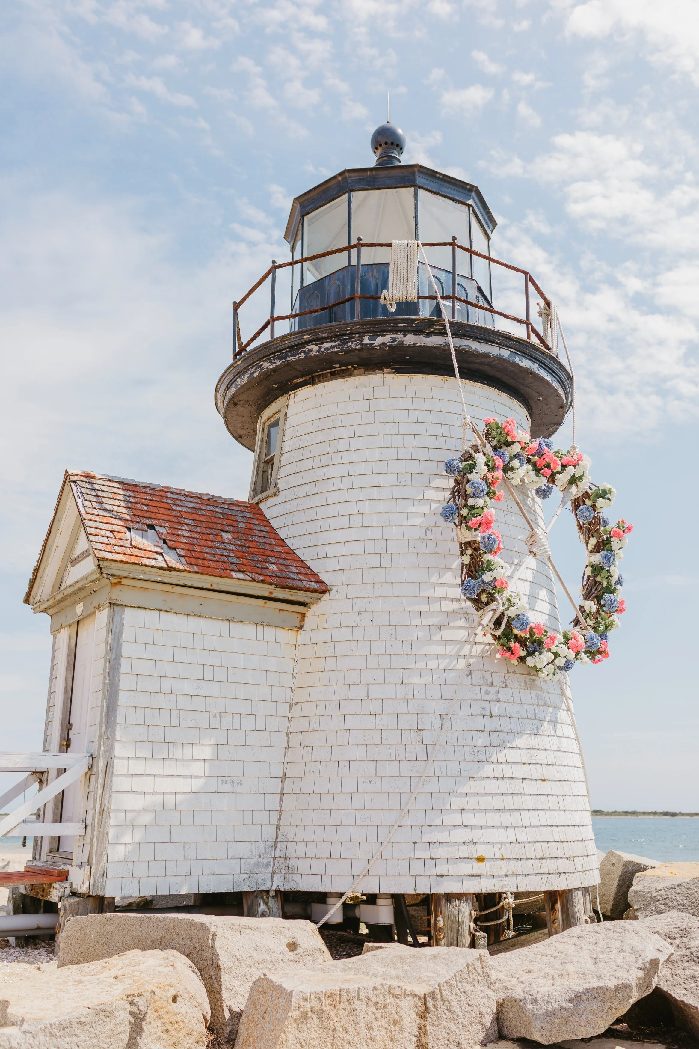 White lighthouse with a red shingled roof, surrounded by a floral wreath, and positioned on a rocky shore under a partly cloudy sky.