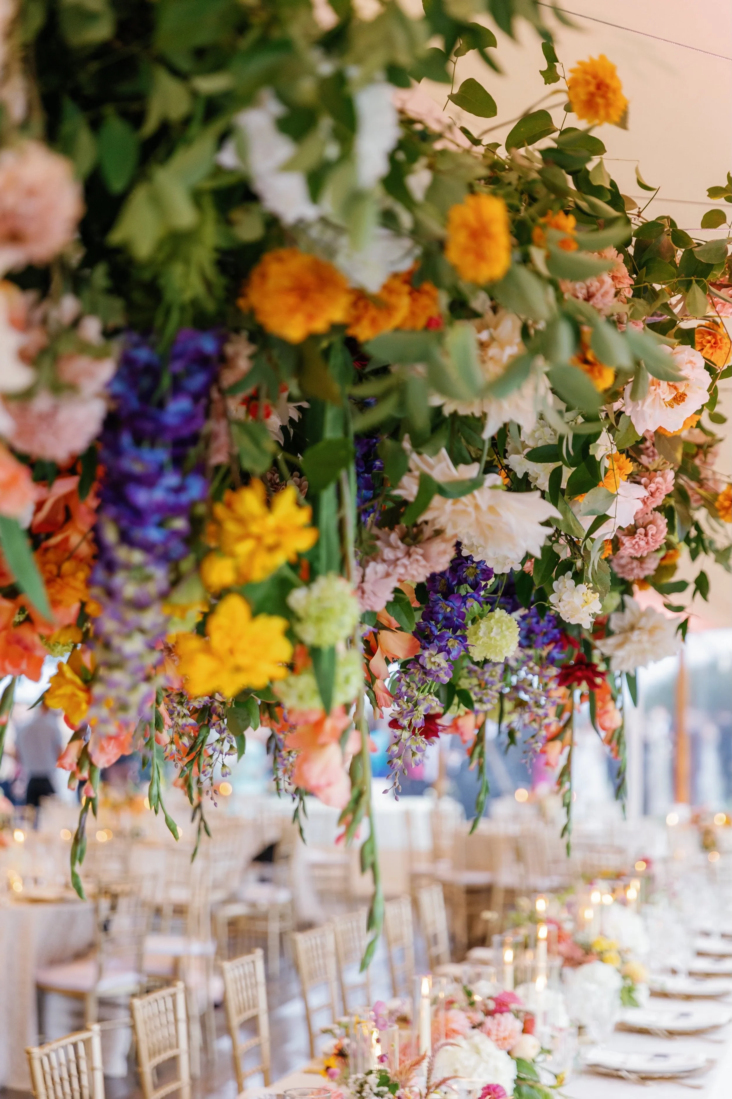 Hanging floral arrangement of yellow, orange, white, purple, and pink flowers over a banquet table set for a celebration.