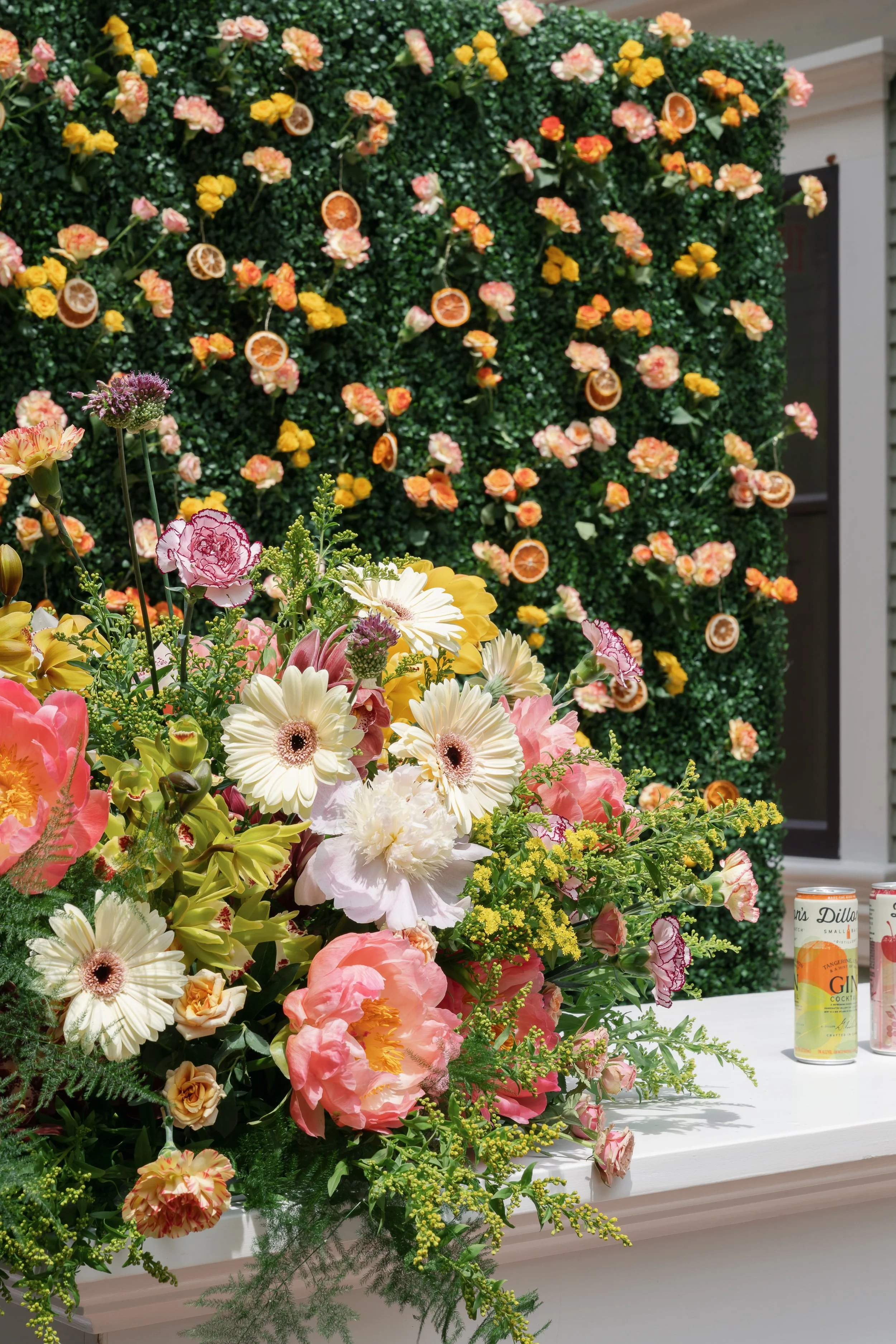 A large floral arrangement with pink, yellow, and white flowers, including carnations, daisies, and roses, placed on a white window sill. In the background, a wall of greenery decorated with orange and pink flowers and slices of dried oranges is visi