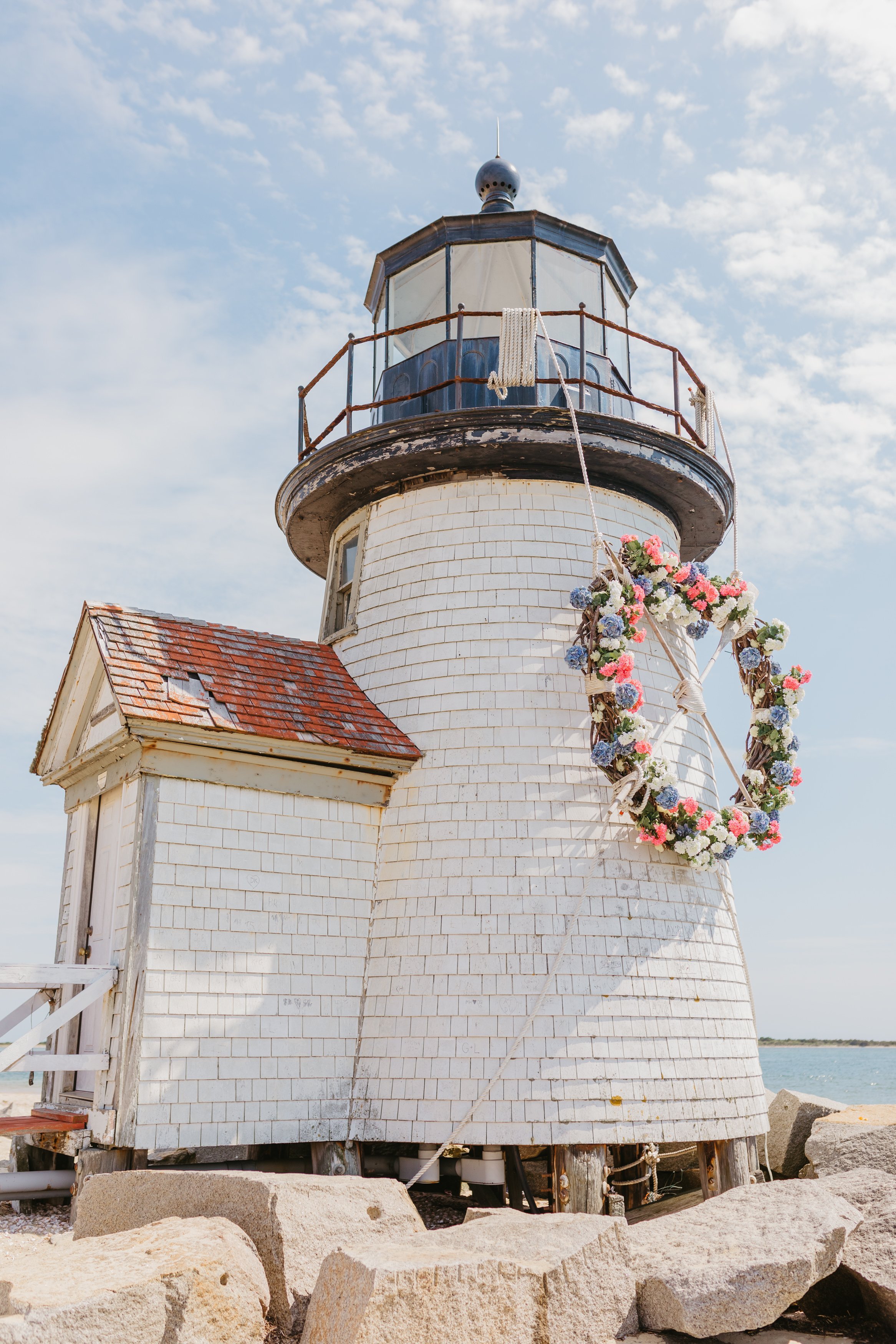 White wooden lighthouse with a lantern room at the top, decorated with a flower wreath hanging on a rope, situated on rocks near the water under a partly cloudy sky.