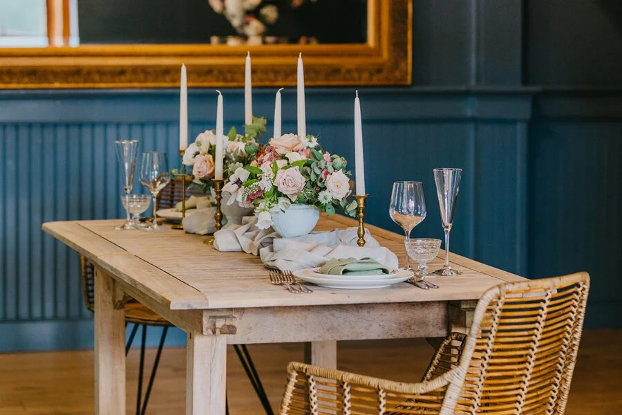 A wooden dining table decorated with a floral centerpiece, tall candlesticks, glassware, and napkins set for a meal, in a room with blue paneled walls and a mirror in the background.