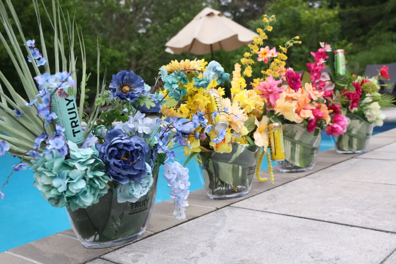 Colorful artificial flower arrangements in glass containers with beer cans among the flowers, set up outdoors near a pool with a brown umbrella in the background.