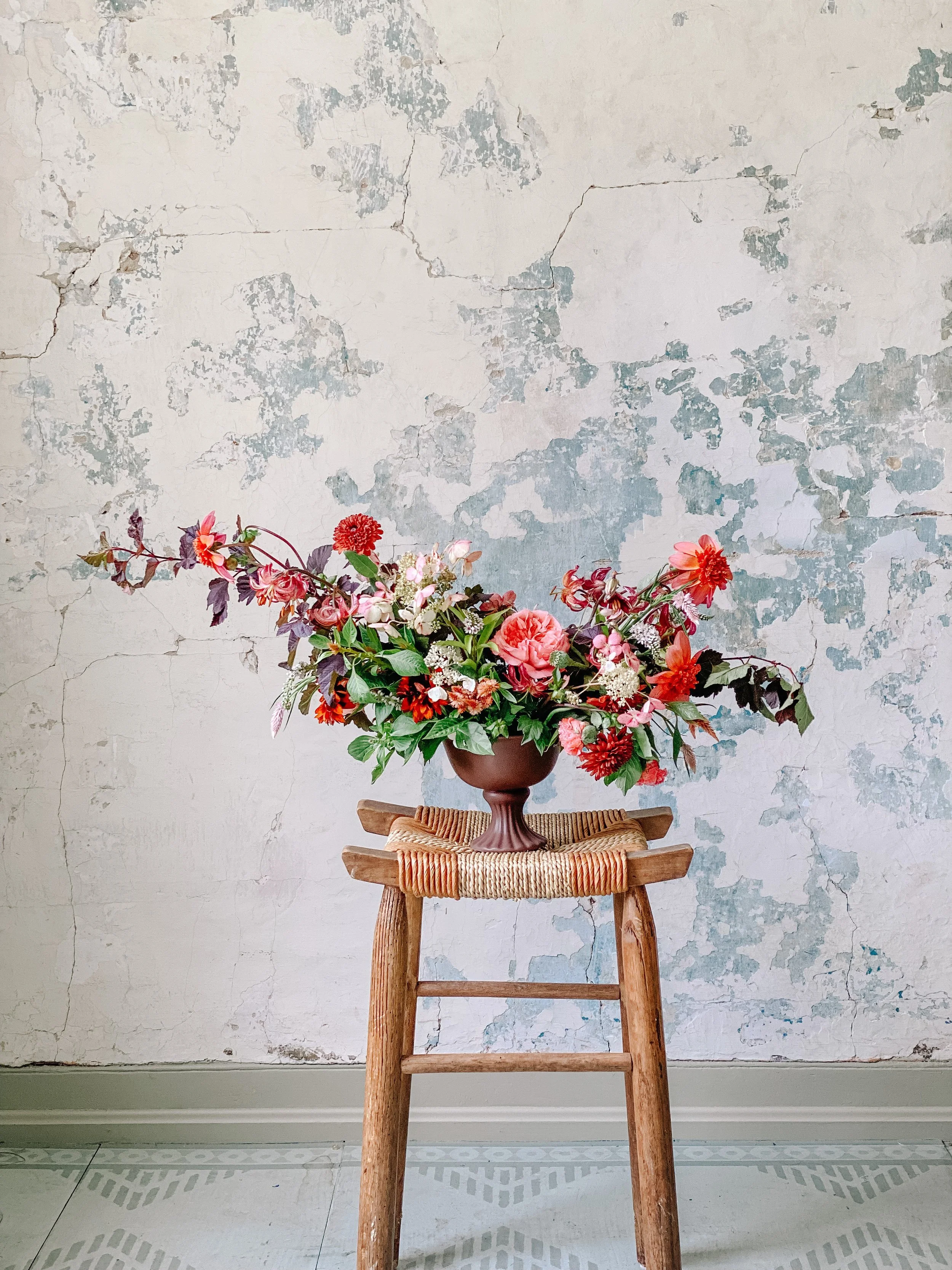A bouquet of pink, red, and purple flowers in a brown ceramic vase on a wooden stool against a distressed white wall background.