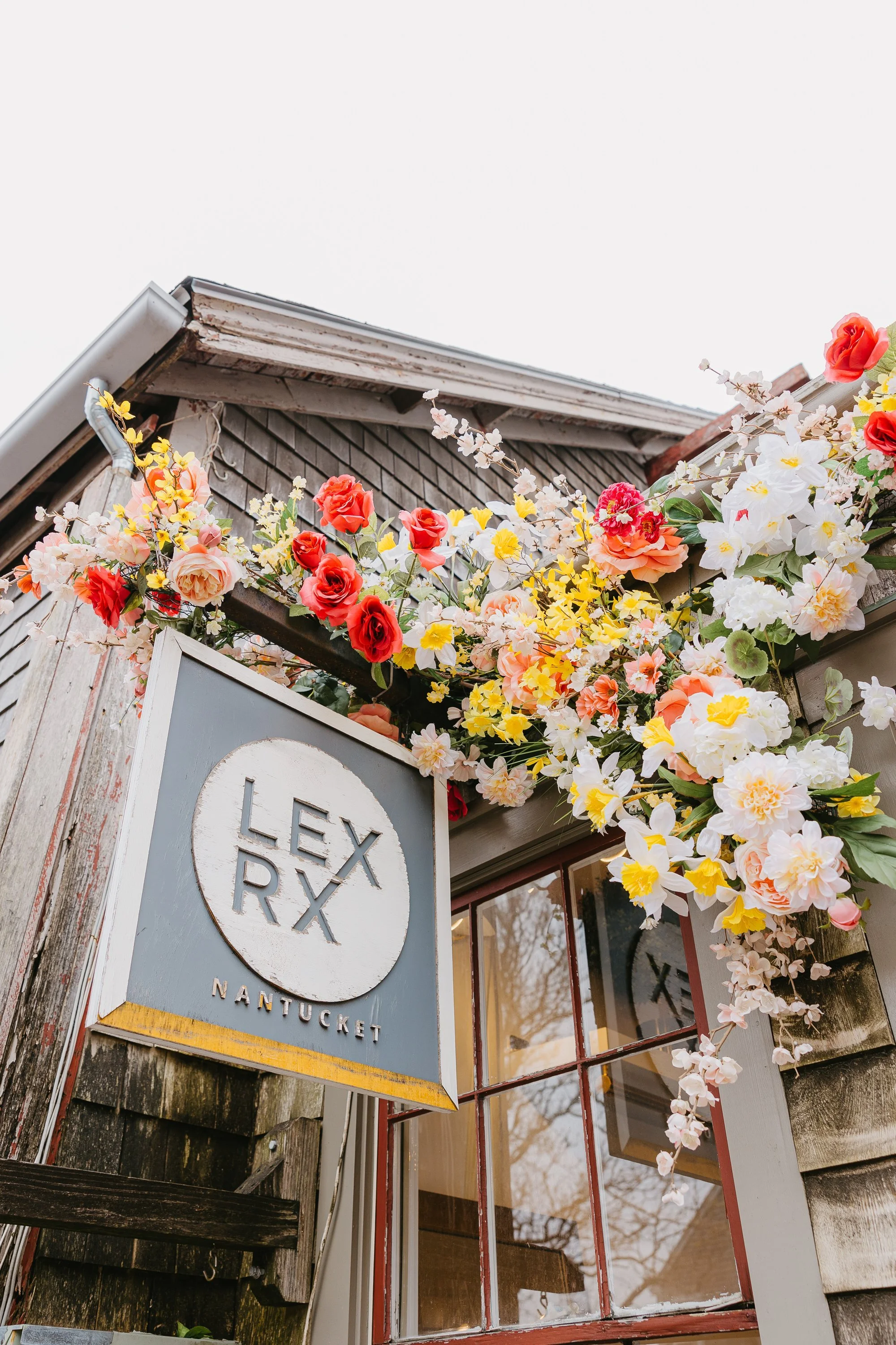 Flower arrangement with white, yellow, pink, and red blooms hanging above a sign that reads "Lex R" with "Nantucket" underneath, attached to a rustic wooden building with a glass window.