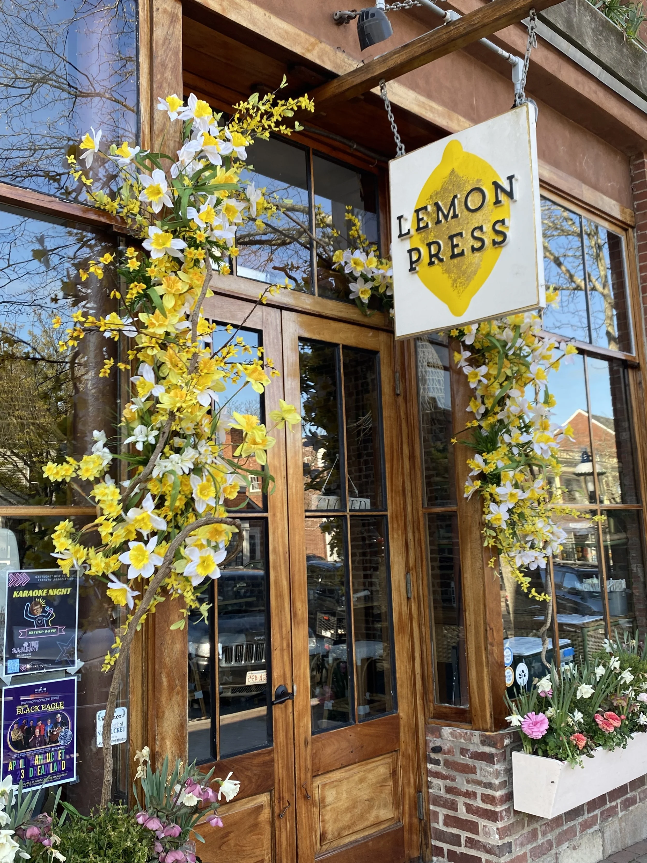 The exterior of a restaurant named Lemon Press with a wooden door, large glass windows, and yellow and white flowers decorating the entrance. A sign hangs above the door with the restaurant's name.
