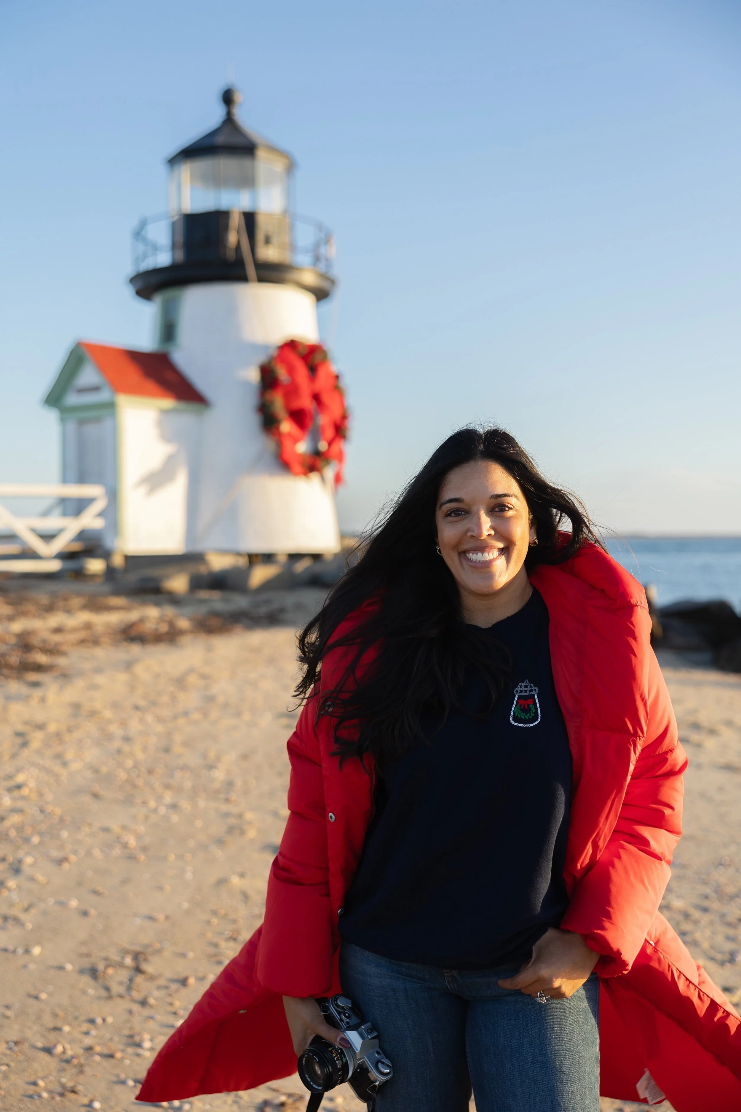 A woman with long dark hair smiling on a beach in front of a lighthouse decorated with a red wreath, holding a camera in her hand, wearing a red jacket and dark shirt with a patch, during sunset or sunrise.