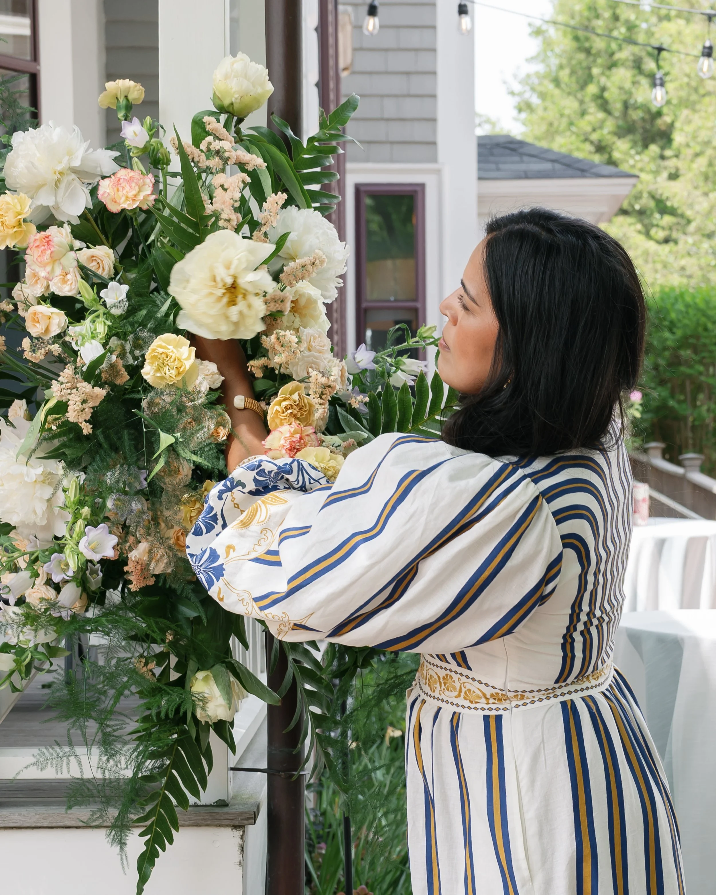 Woman with dark hair arranging a large bouquet of white, cream, and pastel pink flowers on a porch, with green foliage and a gray house in the background.