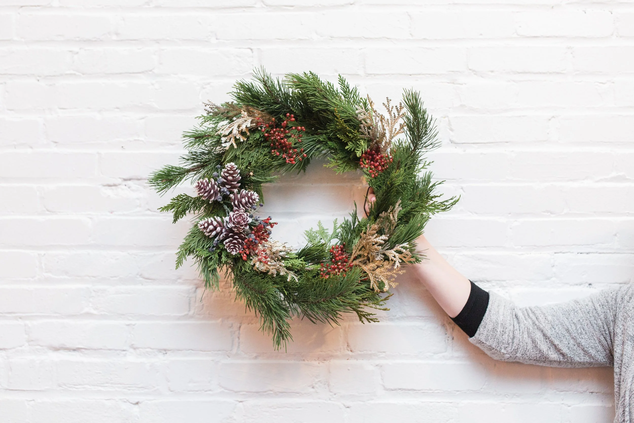 A person holding a Christmas wreath decorated with pine branches, pinecones, red berries, and gold accents against a white brick wall.