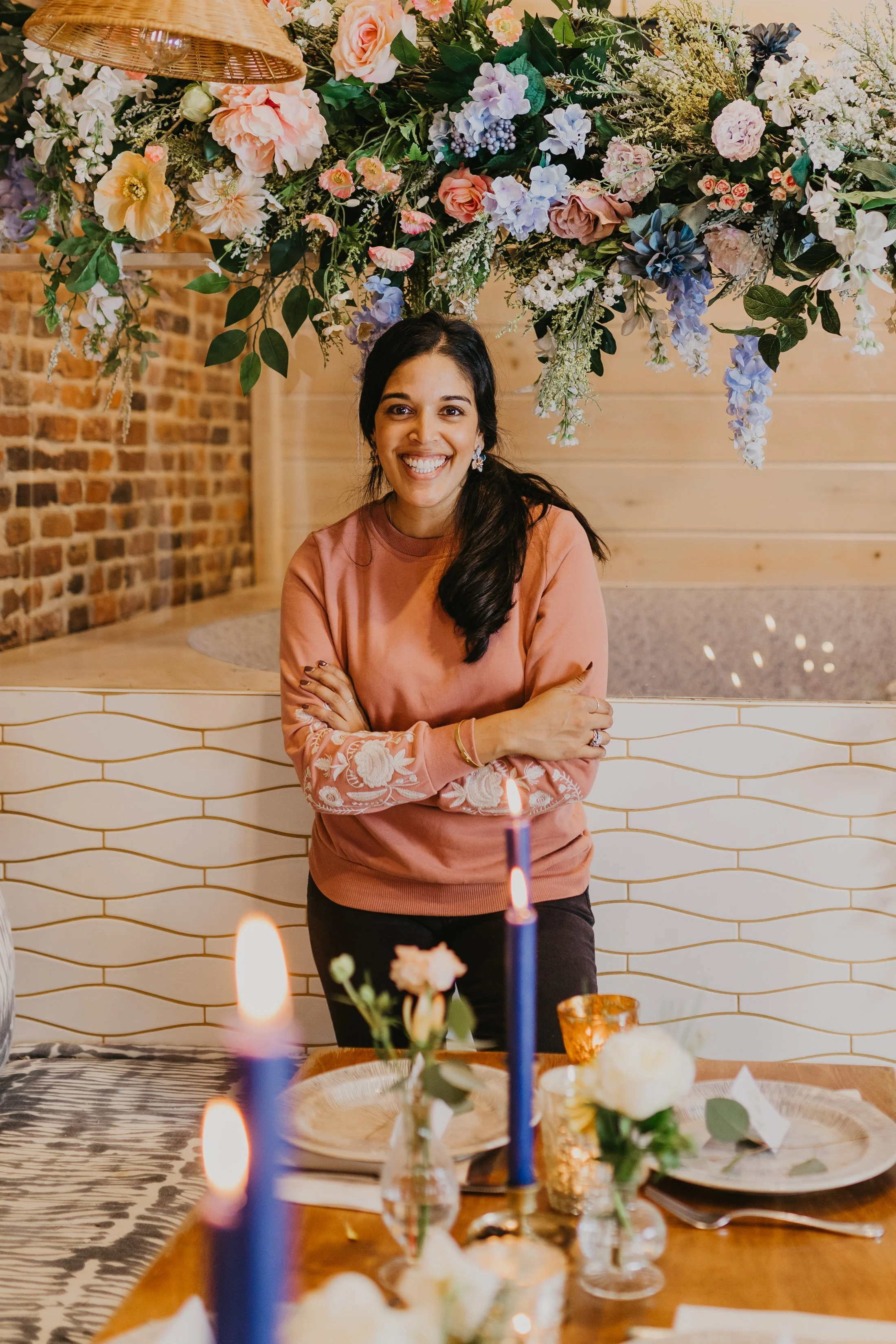 A woman sitting at a decorated table with lit candles, flowers, and a birthday cake, smiling in a festive setting with a large flower arrangement overhead and a brick wall background.