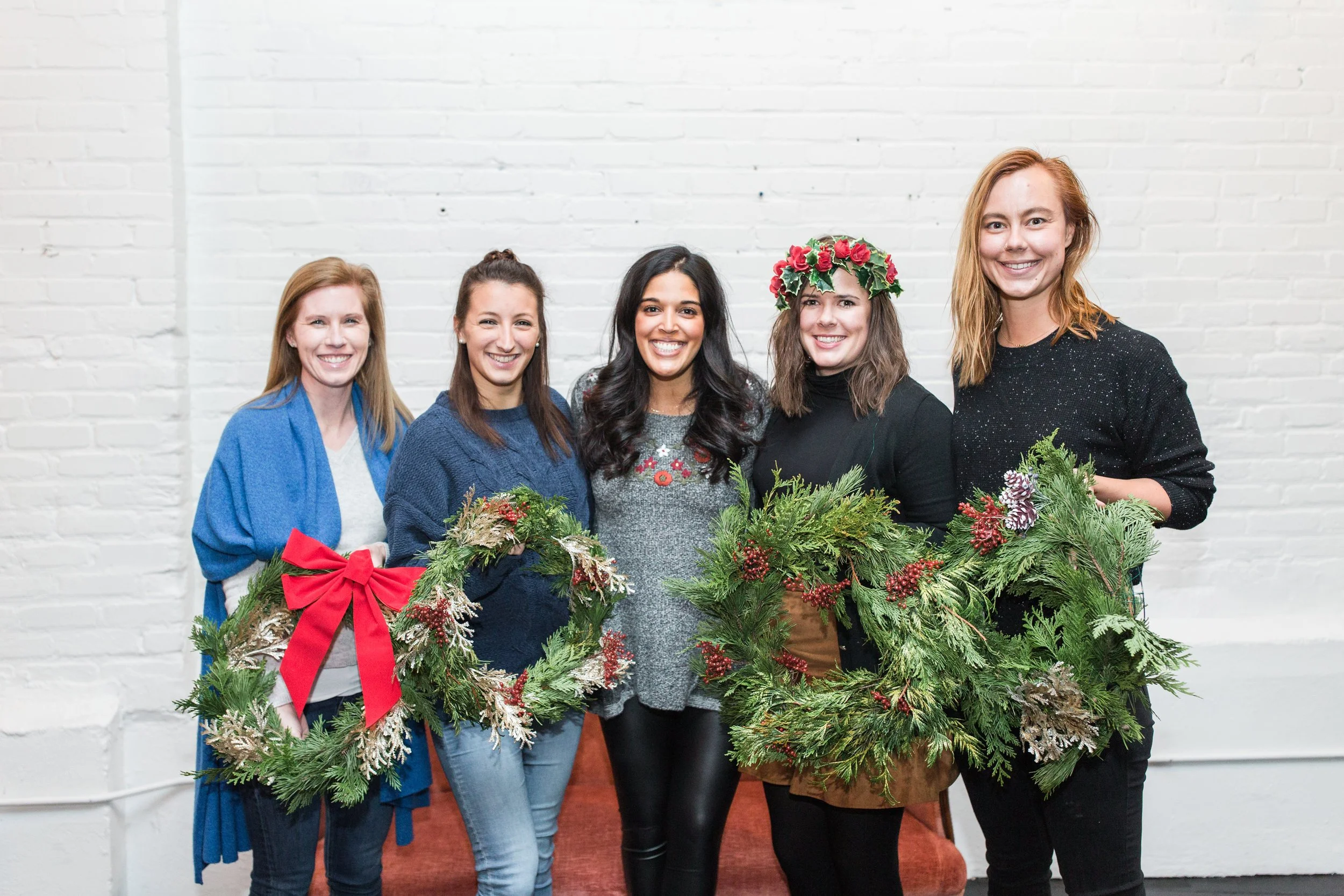 A group of five women standing together, smiling, holding Christmas wreaths and decorated with holiday attire, set against a white brick wall.