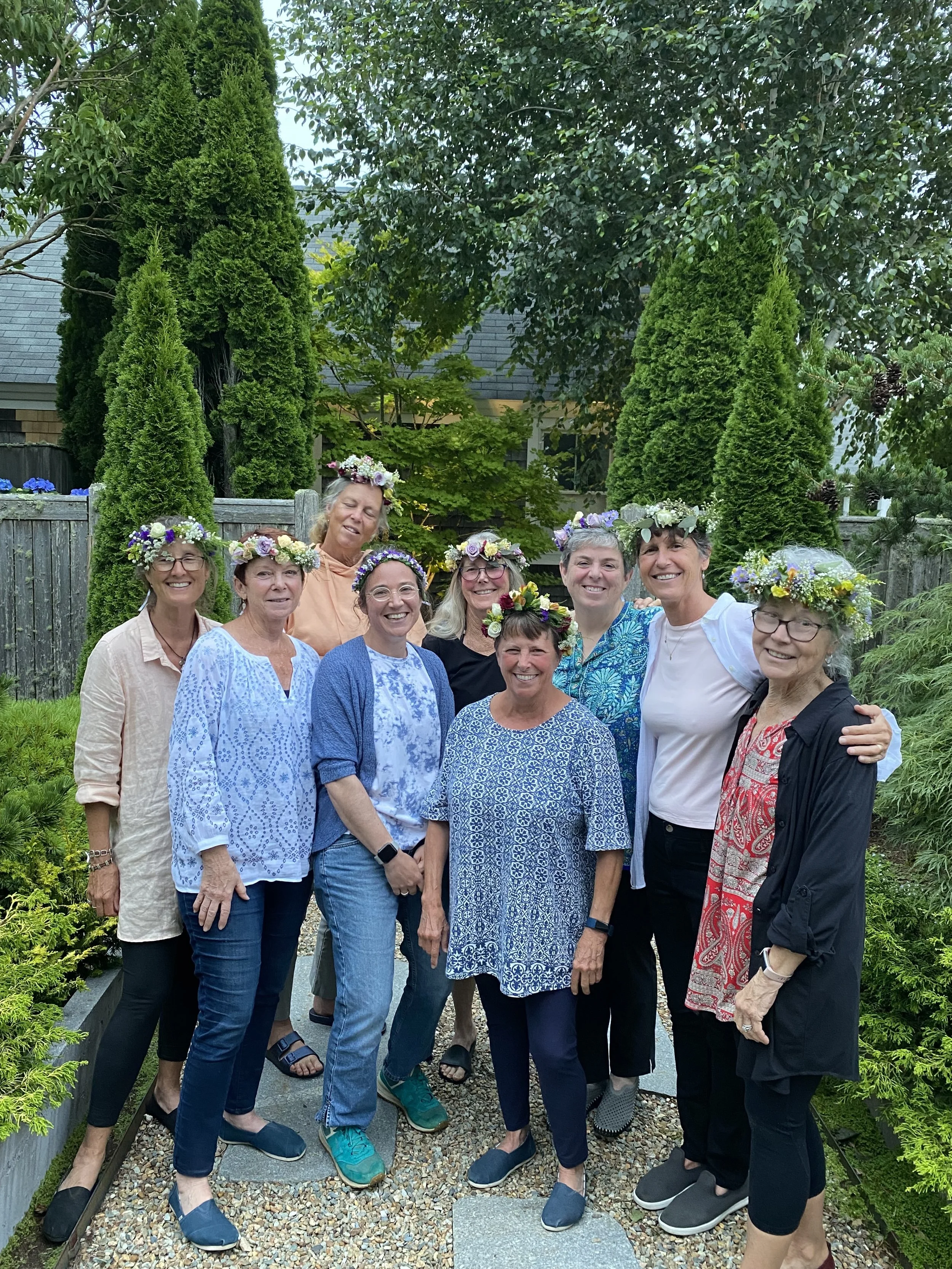 A group of women standing outdoors in a garden, wearing flower crowns, smiling, and posing for a photo.