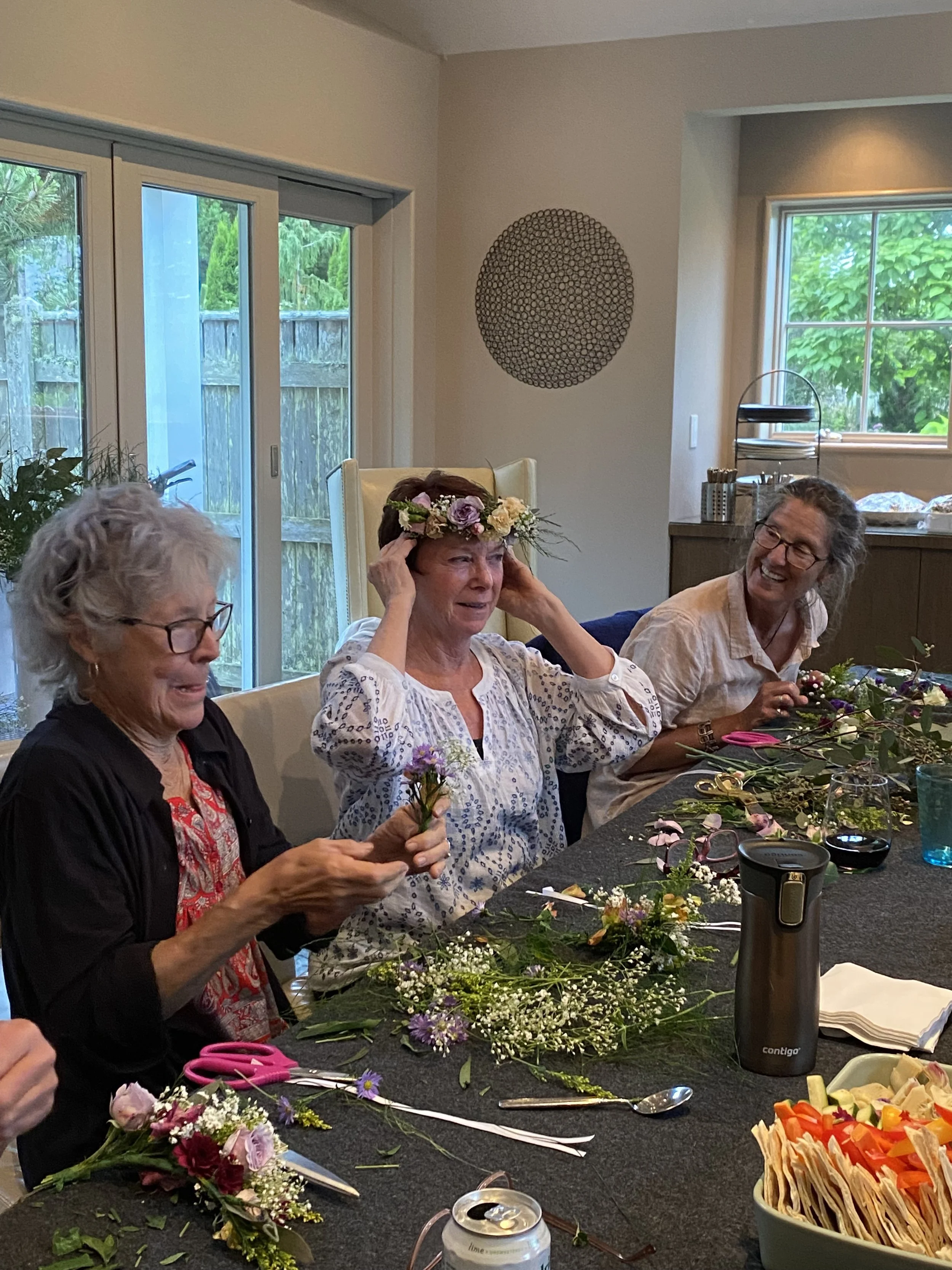 Older women sitting at a table making floral arrangements with various flowers, scissors, and tools, while smiling and chatting in a bright room with large windows.
