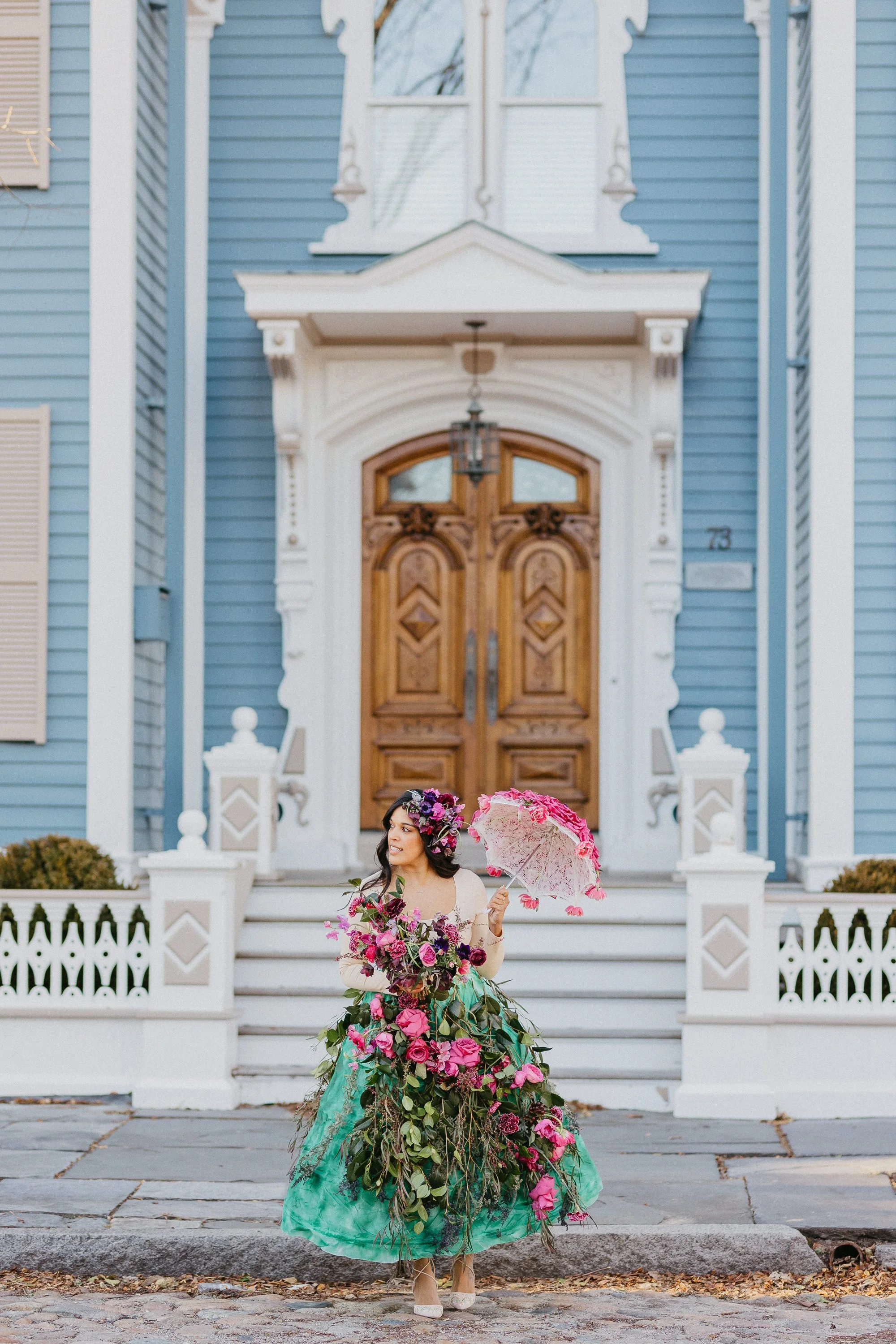 Woman in colorful floral dress holding a pink umbrella standing in front of a blue house with a wooden door and white porch.