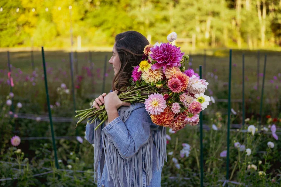 Woman with long brown hair holding a large bouquet of colorful flowers with a garden or flower field in the background.