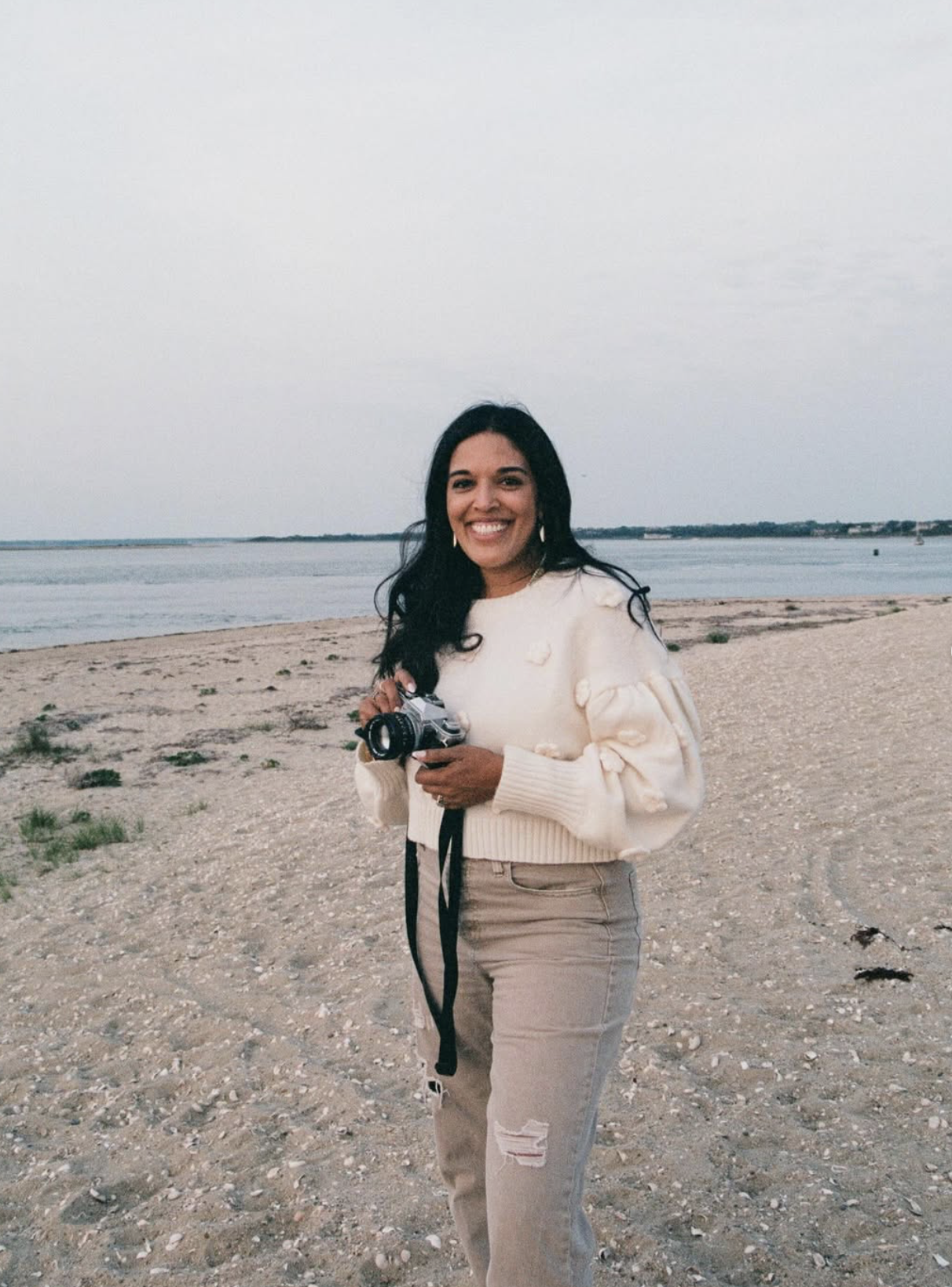 A woman with long black hair holding a camera on a beach with water and a cloudy sky in the background.