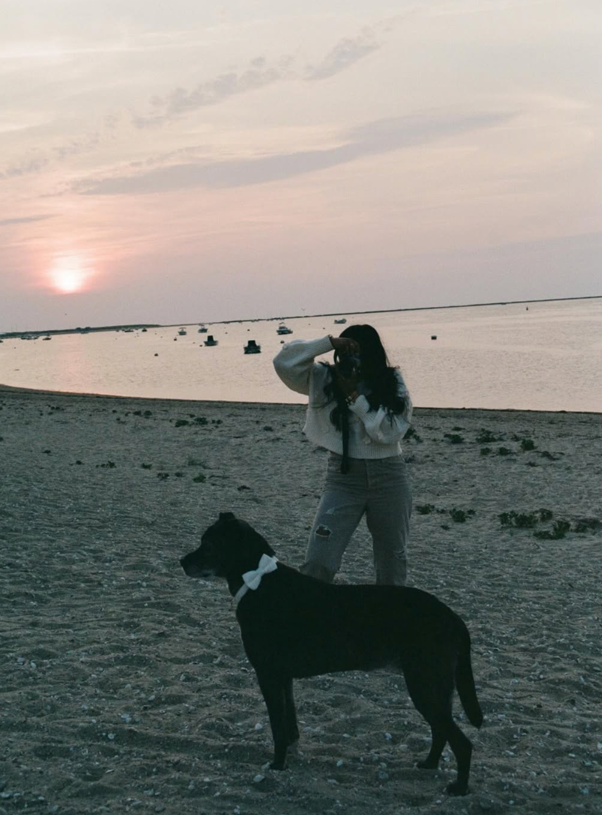 A woman taking a photo on a beach during sunset, with a black dog with a white bow in the foreground and boats on the water in the background.