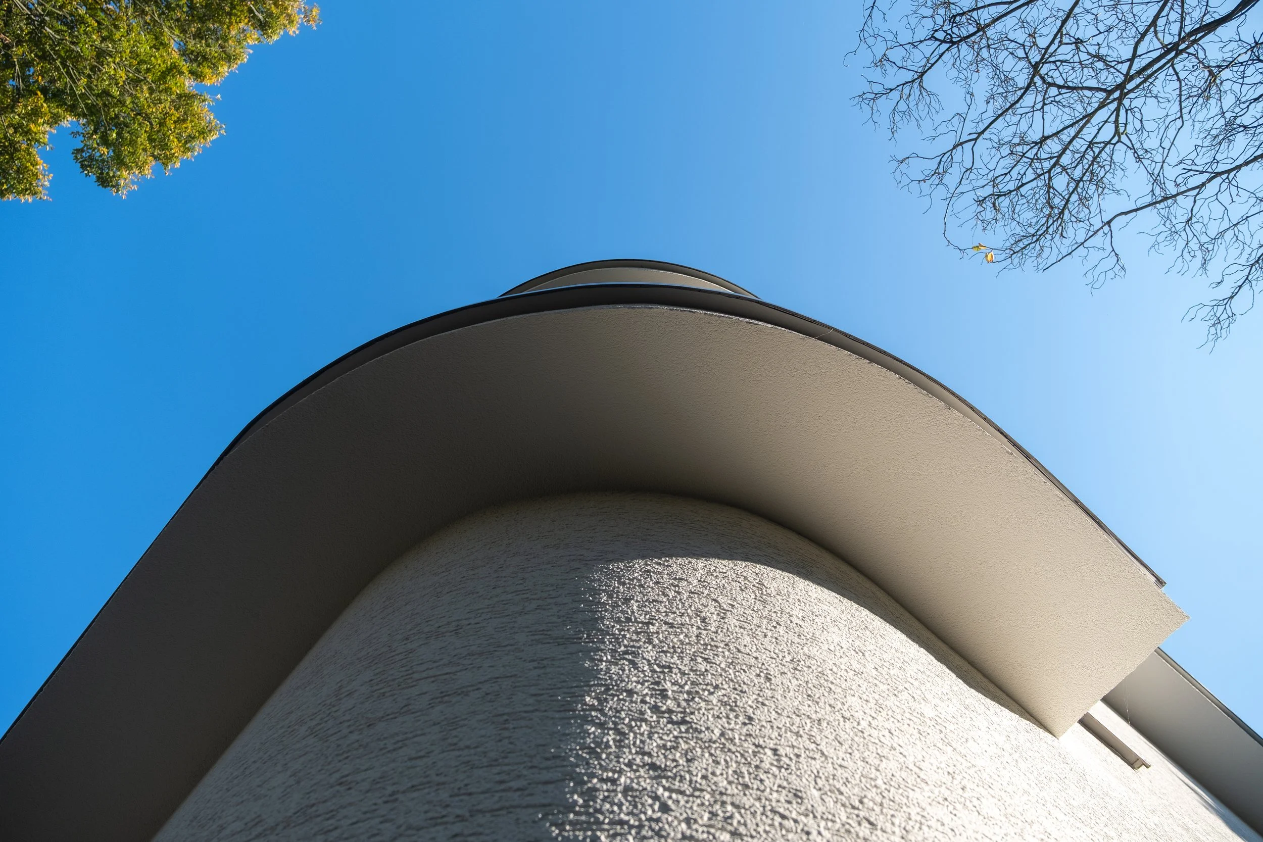 Een moderne witte woning met een ronde, uitkragende dakrand, gezien vanaf onderen tegen een blauwe lucht met een boom met groene bladeren en een boom met takken zonder bladeren.