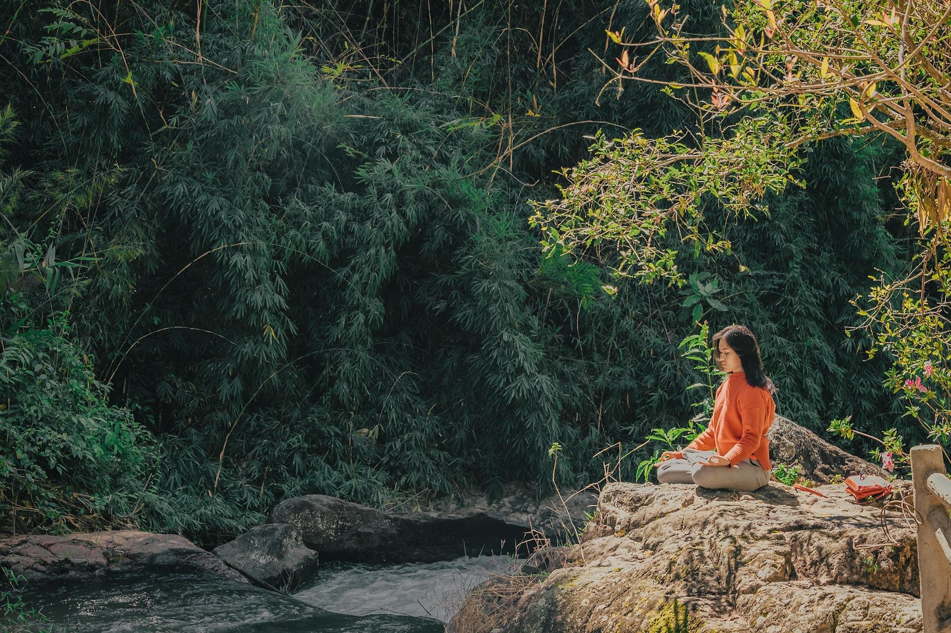 A woman sitting cross-legged meditating on a rock by a river with lush green foliage in the background.