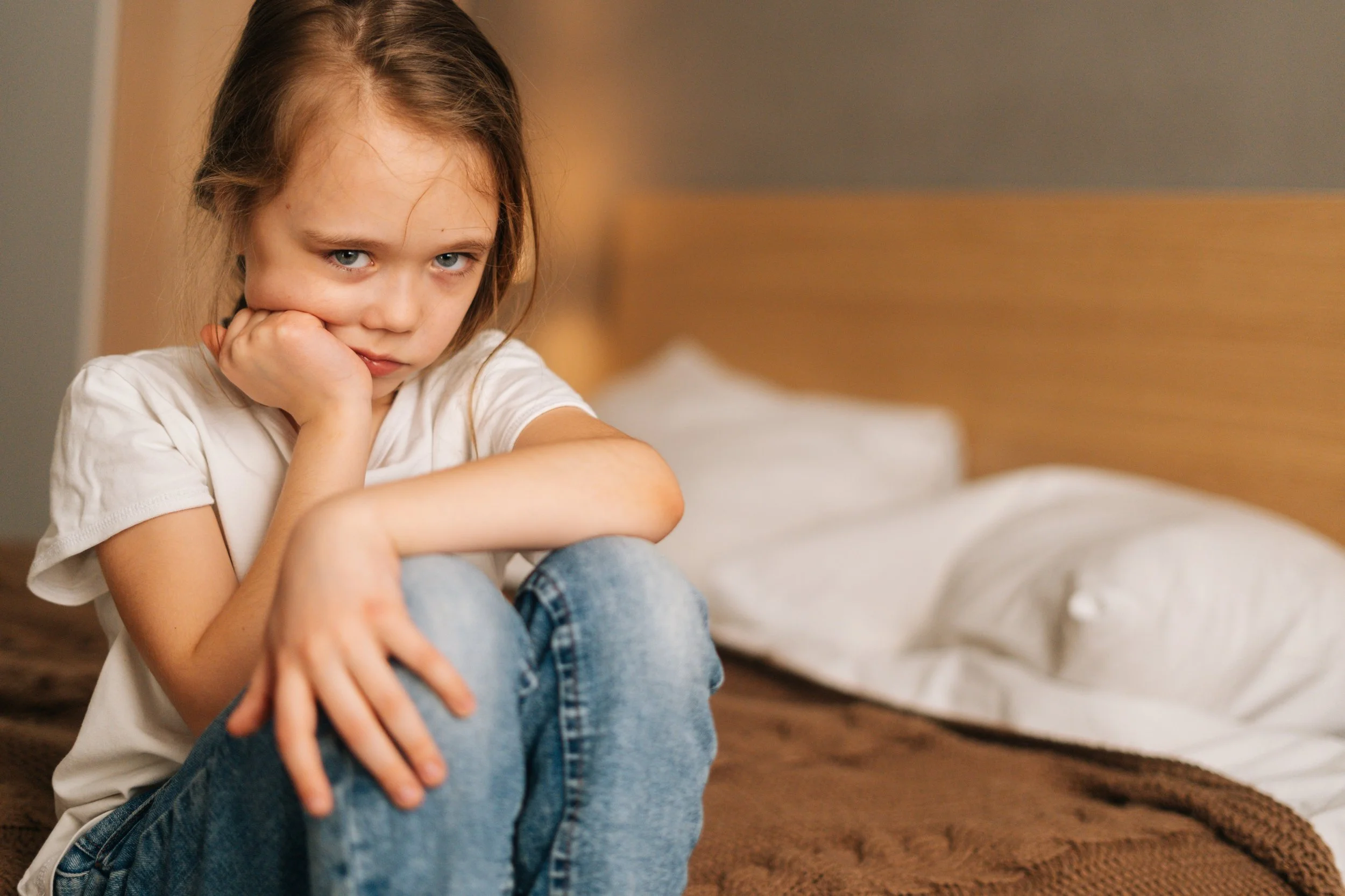 A young girl with brown hair and blue eyes sitting on a bed, appearing upset or bored, with a white shirt and jeans. The bed has a brown blanket and white pillows in the background.