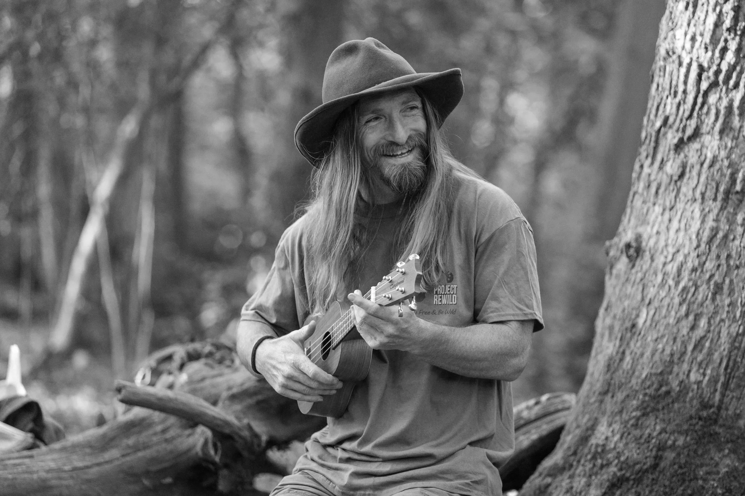 Man with long hair and beard, wearing a hat, sitting near a campfire surrounded by children.