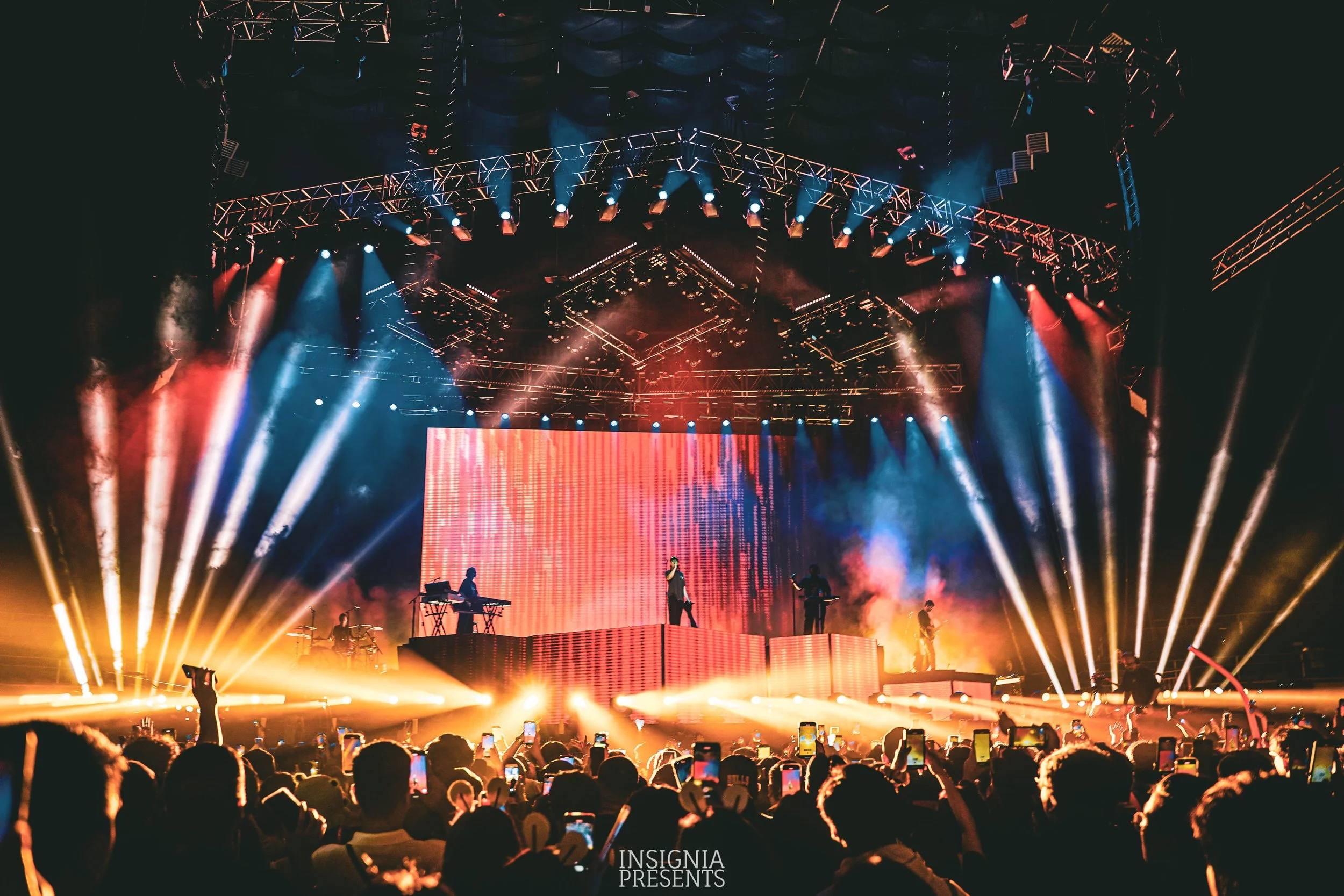A live concert stage with colorful lighting and performers. The stage has a large LED screen, and the audience is taking photos with their phones.