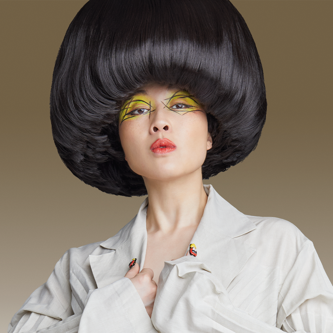 Woman with large, voluminous black hair, wearing a beige shirt with small colorful pins, and vibrant green eyeshadow with artistic black lines, making a confident expression.
