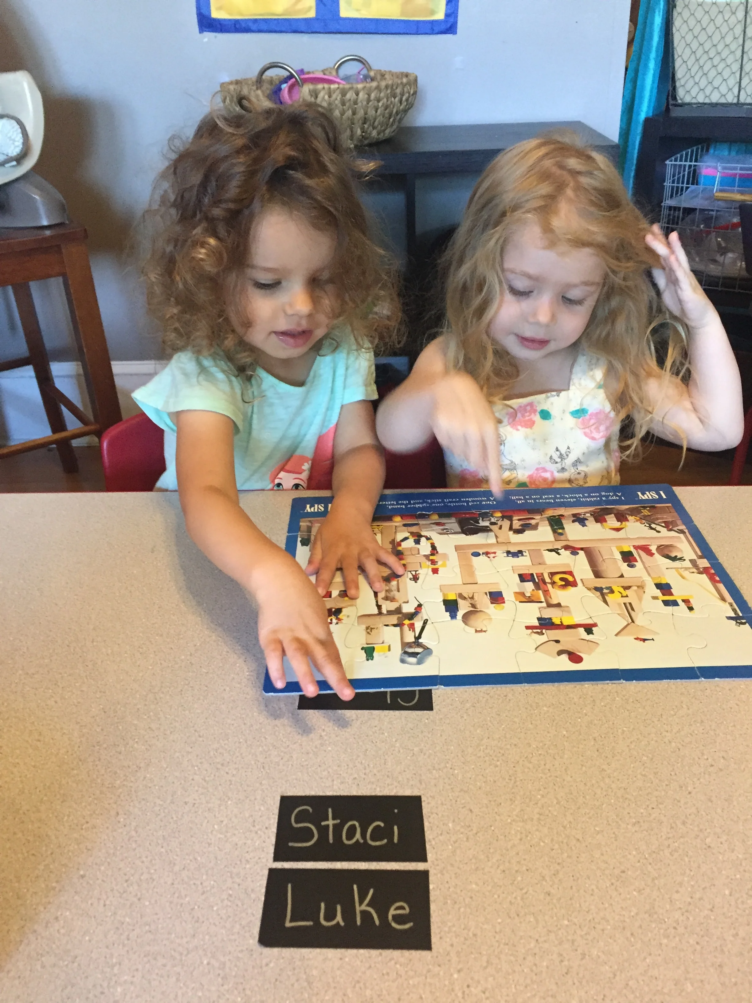 Two young girls with curly hair are playing with a puzzle on a table. There are two black labels on the table that read 'Staci' and 'Luke'.