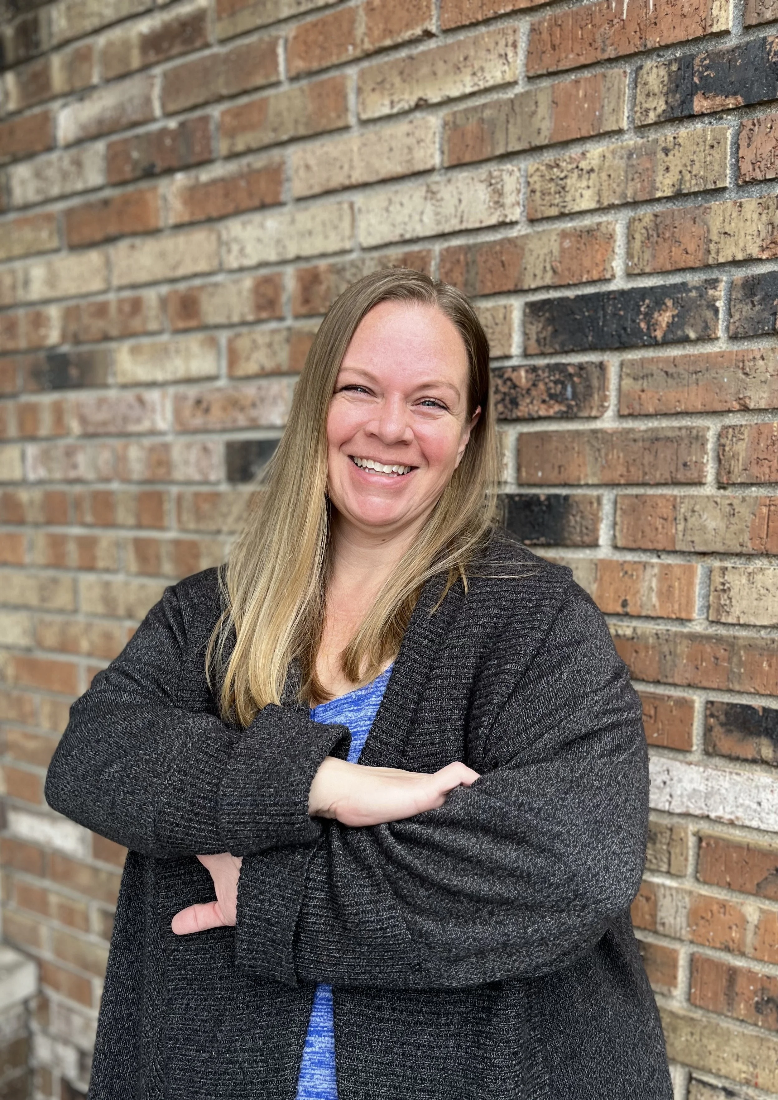 A woman smiling with arms crossed standing against a brick wall.