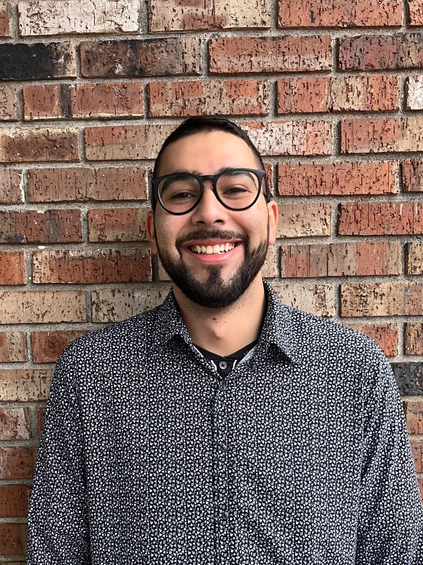 A smiling man with glasses and a beard standing in front of a brick wall.