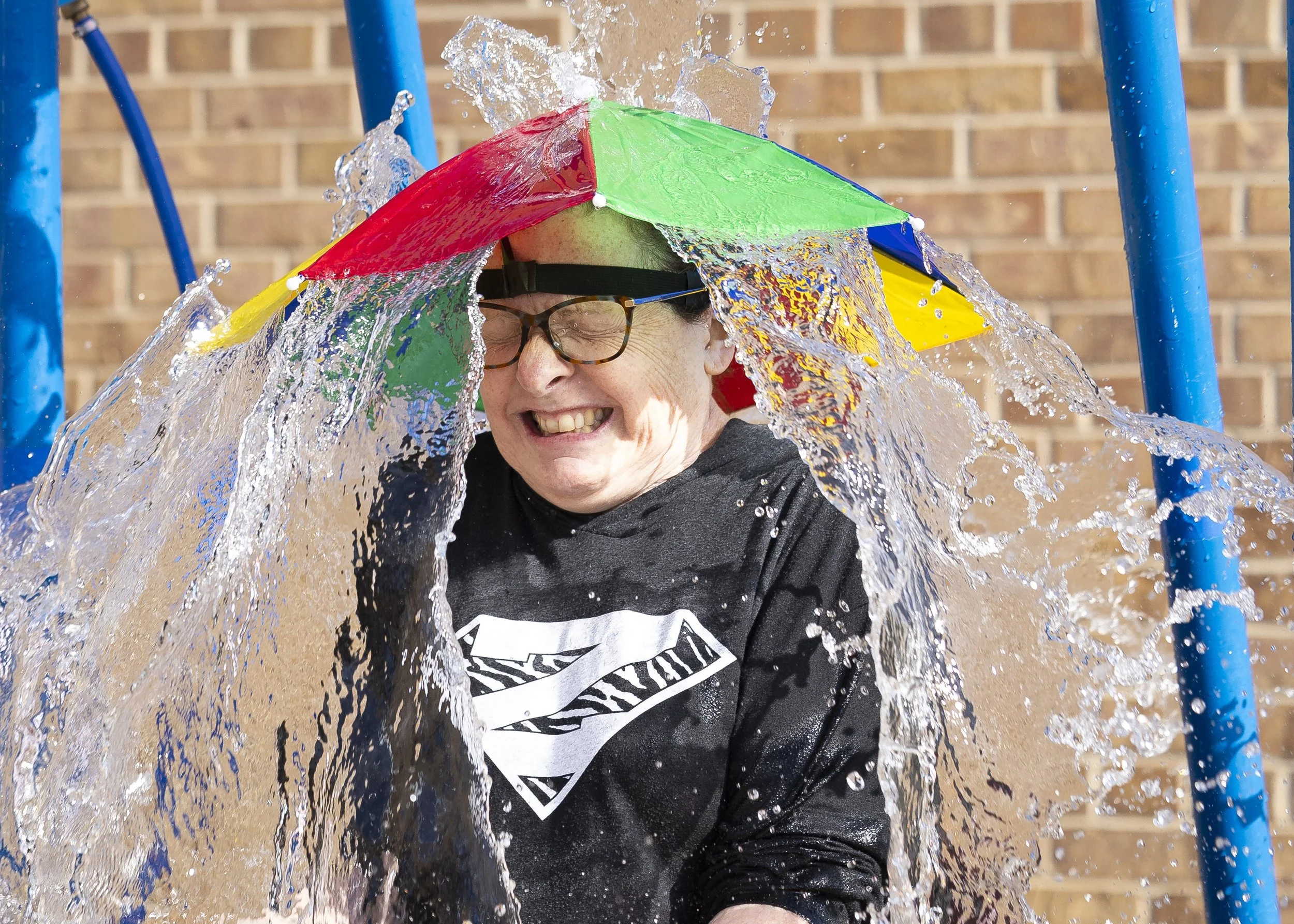  After donning her rainbow umbrella cap, Zeman Elementary School first grade teacher Eileen Schmeichel is doused with water from a splash tower during the Zeman Color Run held on the Zeman Elementary School playground on Saturday, April 13, 2024, in 